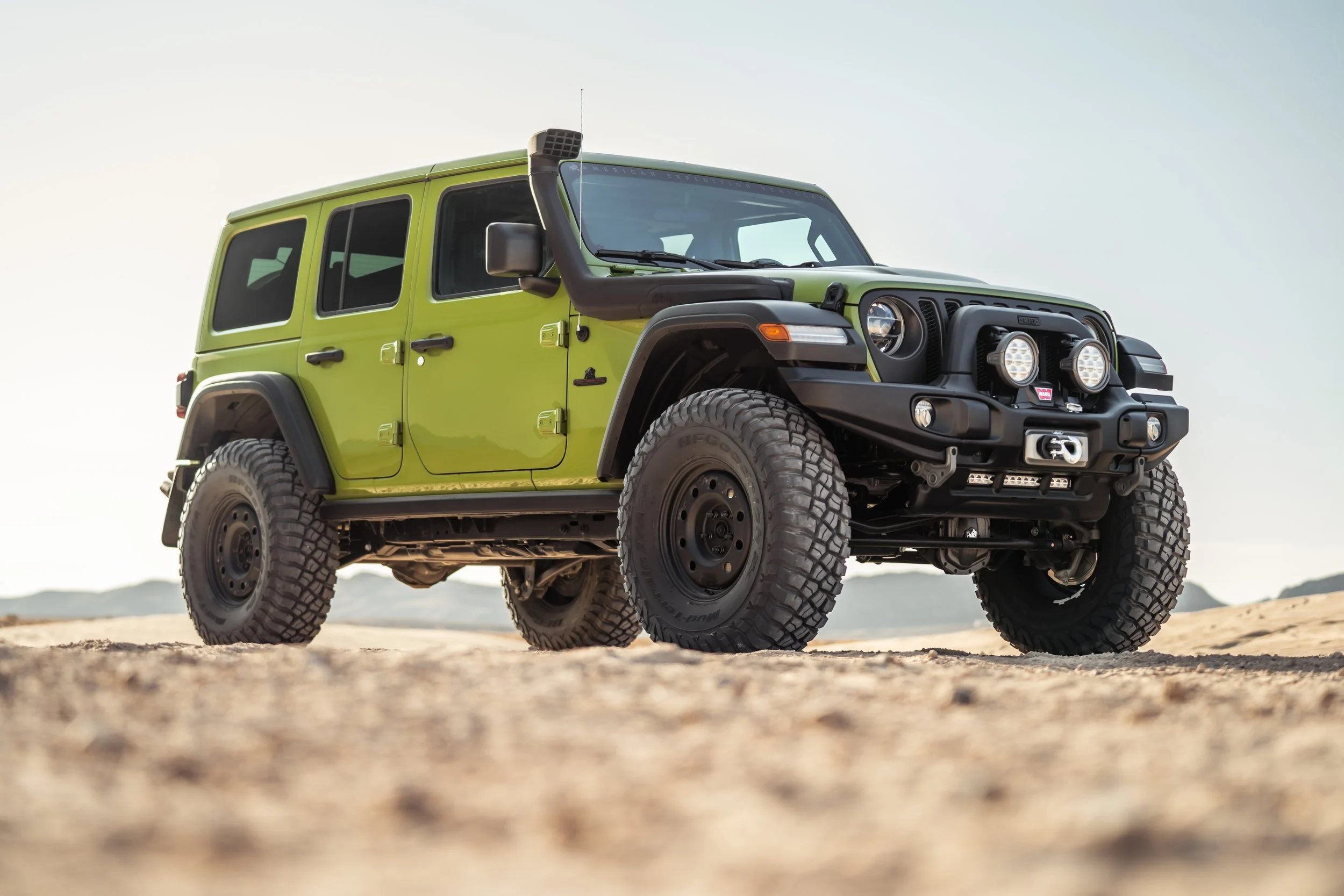 A green off-road SUV parked on sandy terrain with mountains in the background.