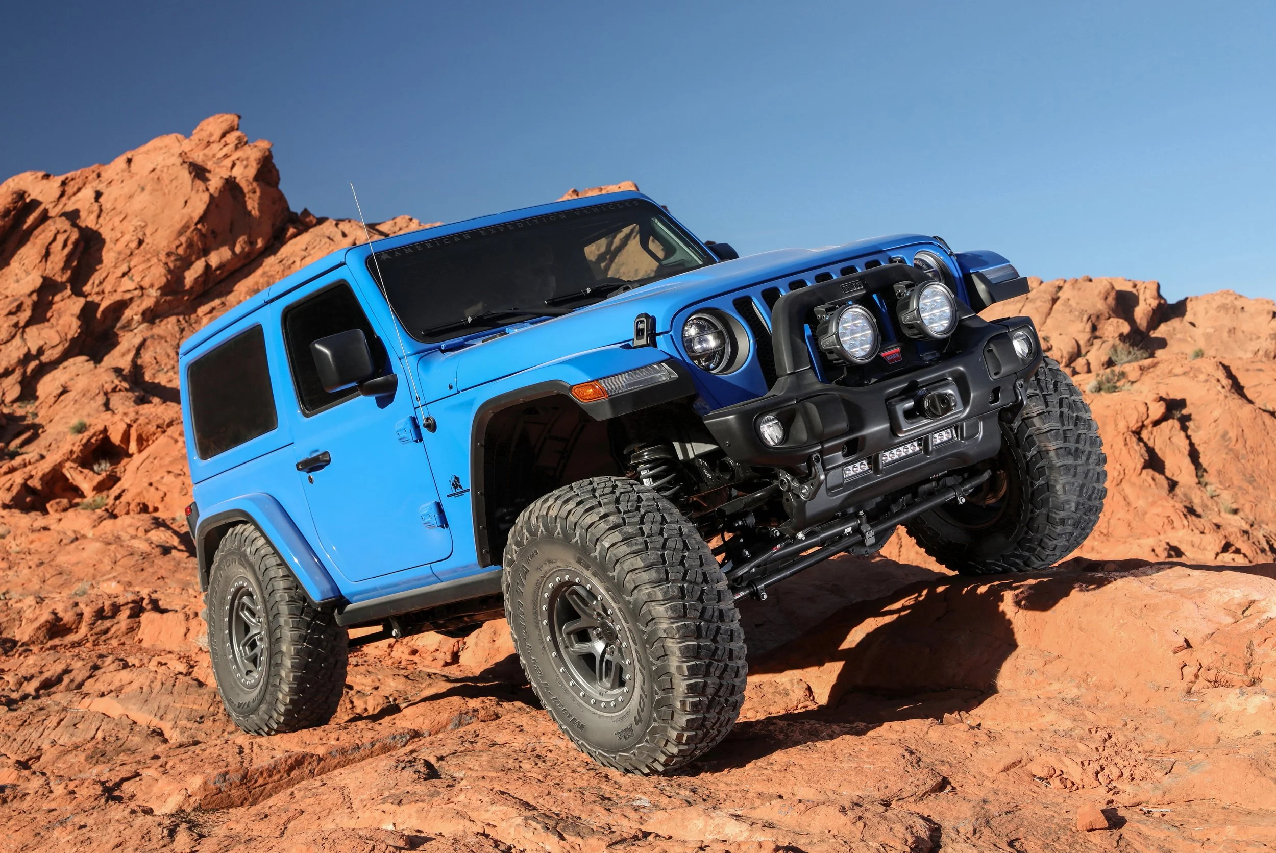 A blue off-road vehicle climbing orange rocky terrain in a desert landscape under a clear blue sky.
