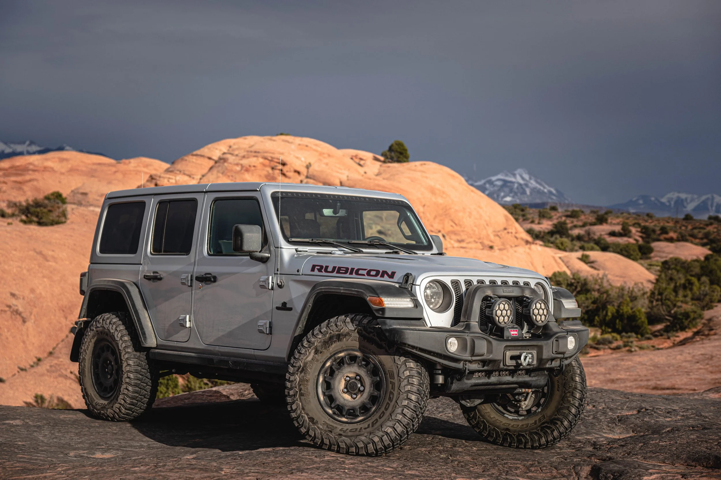 A silver off-road Jeep Rubicon parked on rocky terrain with a desert landscape and mountains in the background.