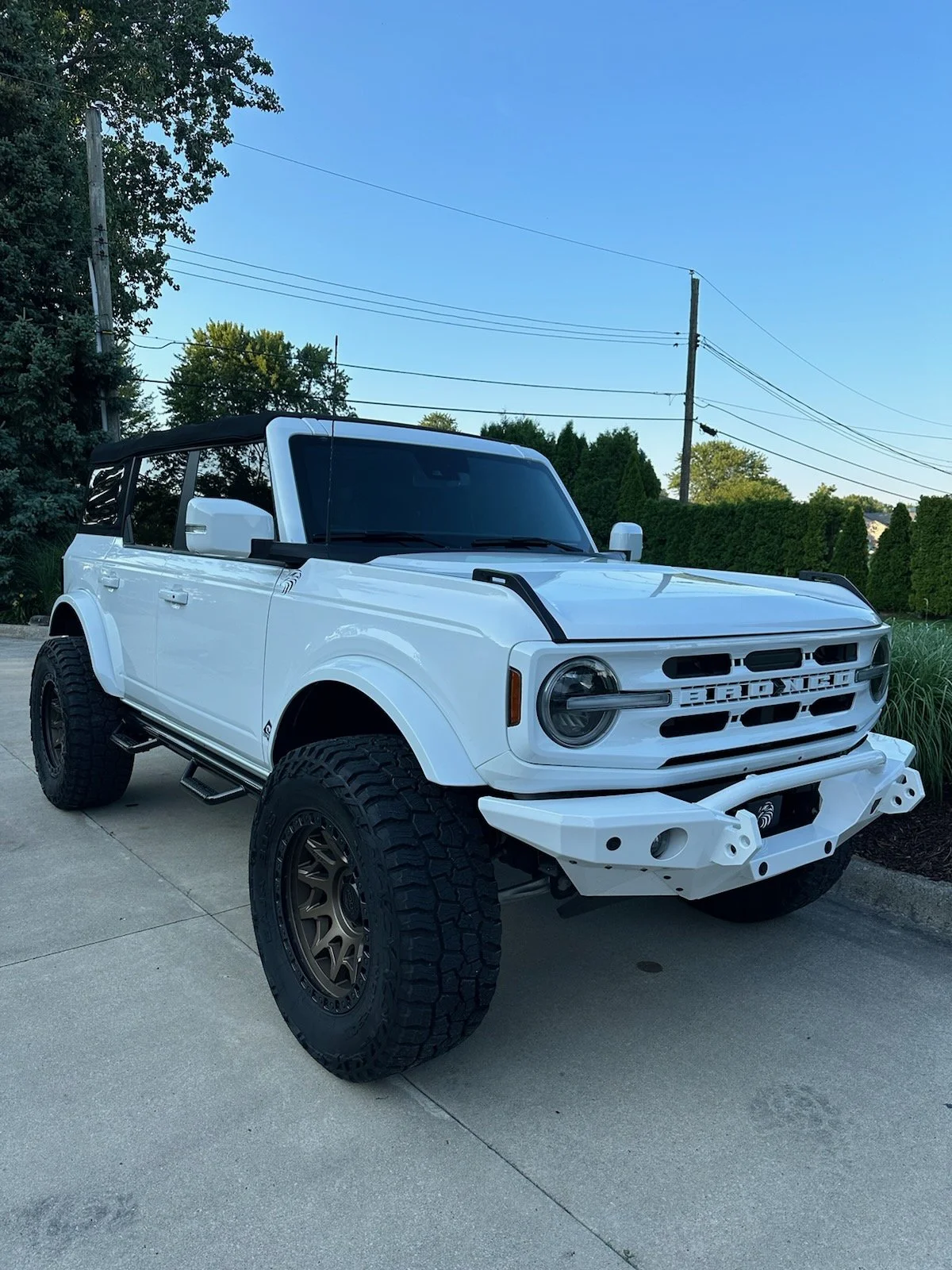 A customized white off-road vehicle with black accents and large tires, parked on a concrete driveway with trees and power lines in the background.