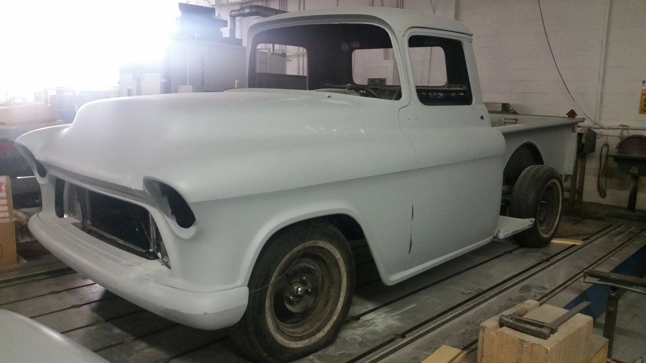 A vintage pickup truck under restoration in a workshop, painted white with black wheels, missing some front parts and windshield, surrounded by tools and equipment.