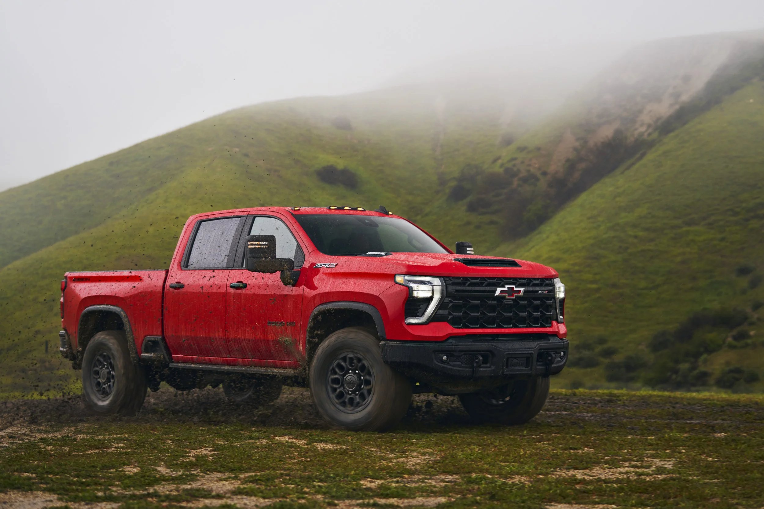 Red Chevrolet Silverado pickup truck driving on a muddy trail in a lush green mountainous landscape with foggy sky