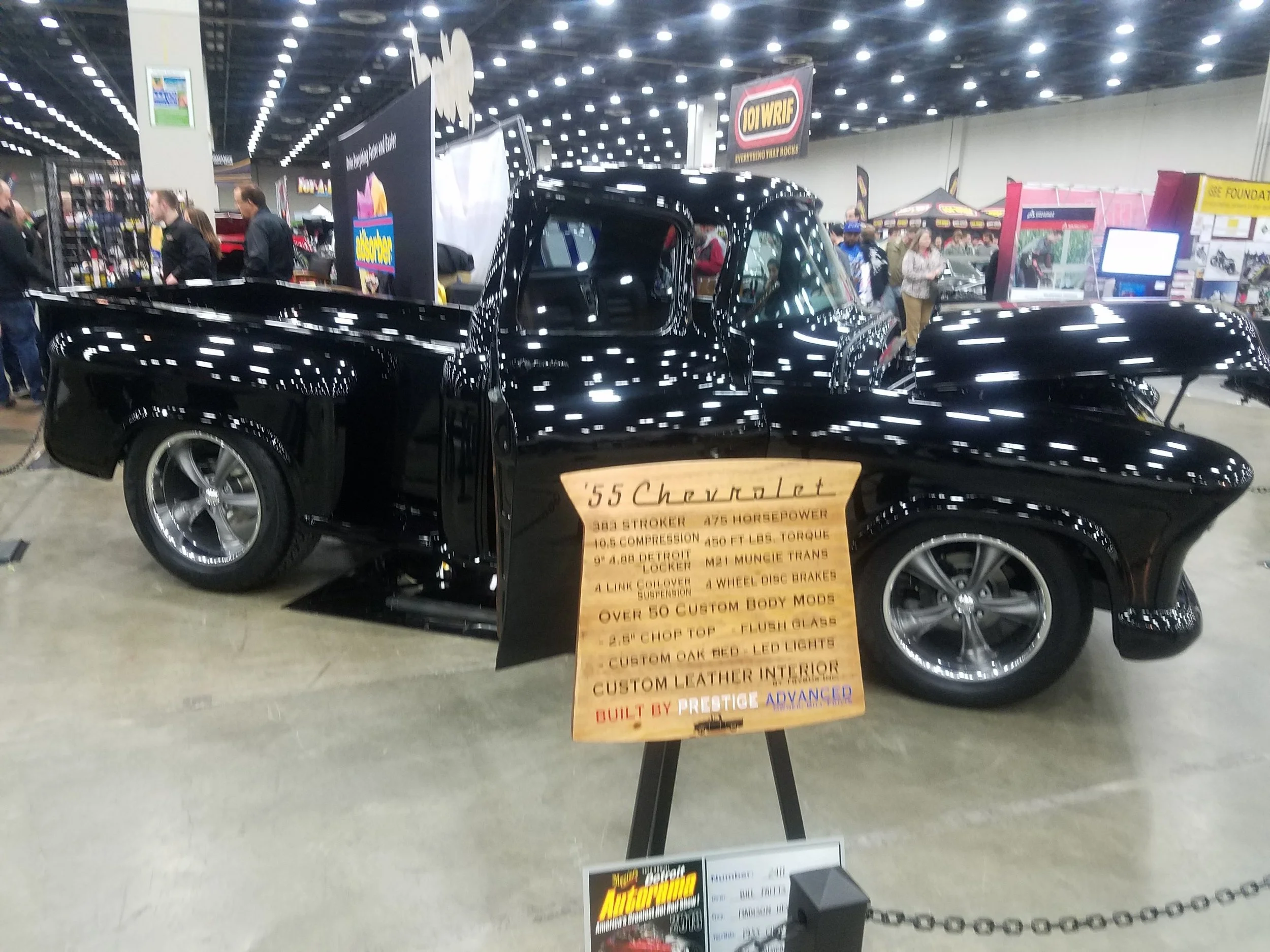 A black 1955 Chevrolet truck on display at a car show with an information sign in front of it, surrounded by other displays and attendees.