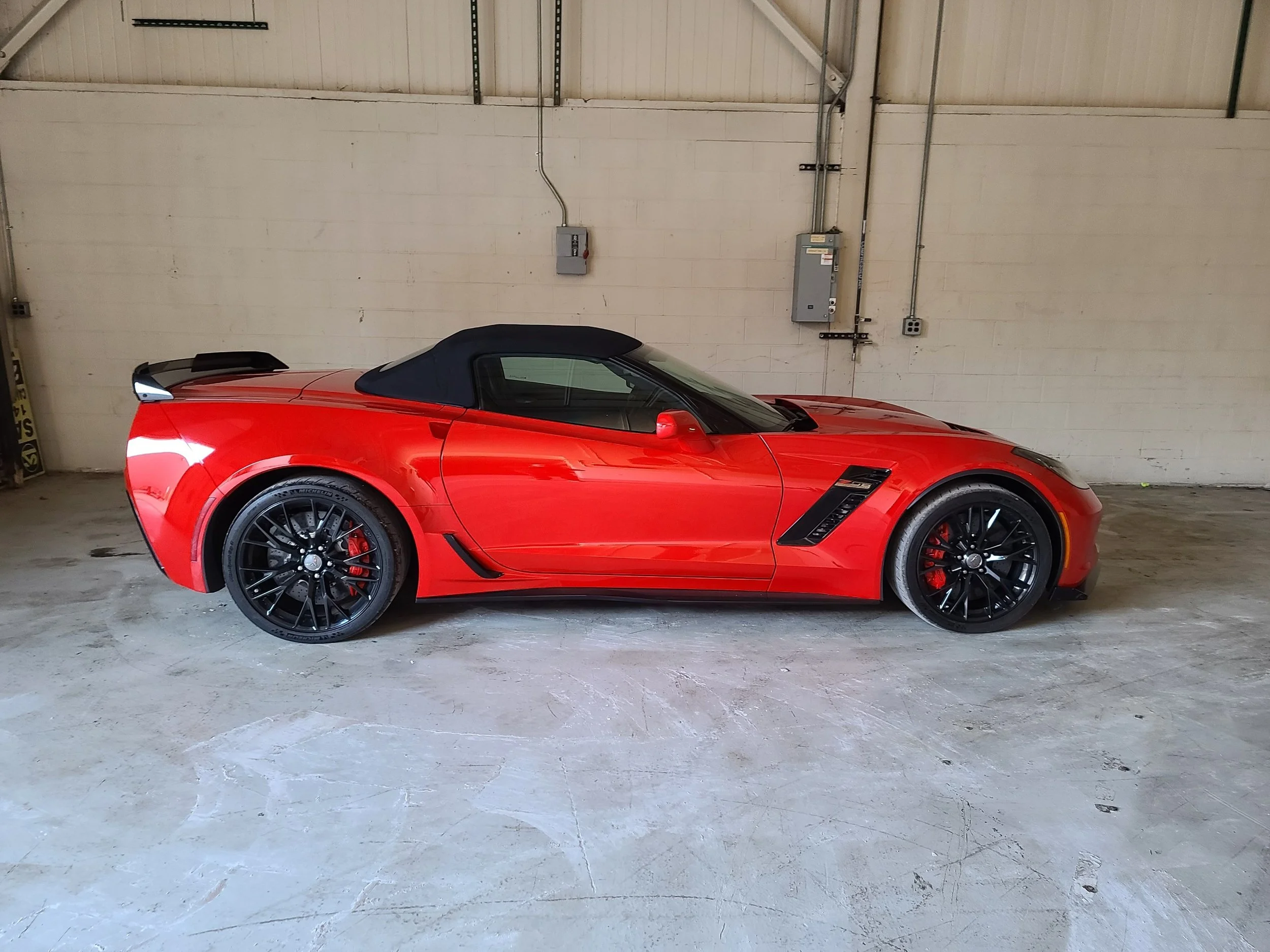 Red Chevrolet Corvette with black wheels parked indoors against a white wall with electrical panels.