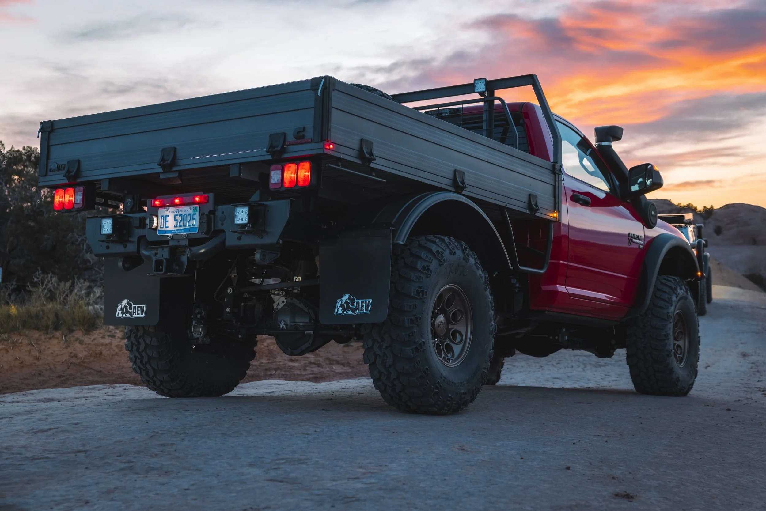 Red and black off-road pickup truck parked on a dirt path at sunset with a mountain landscape in the background.