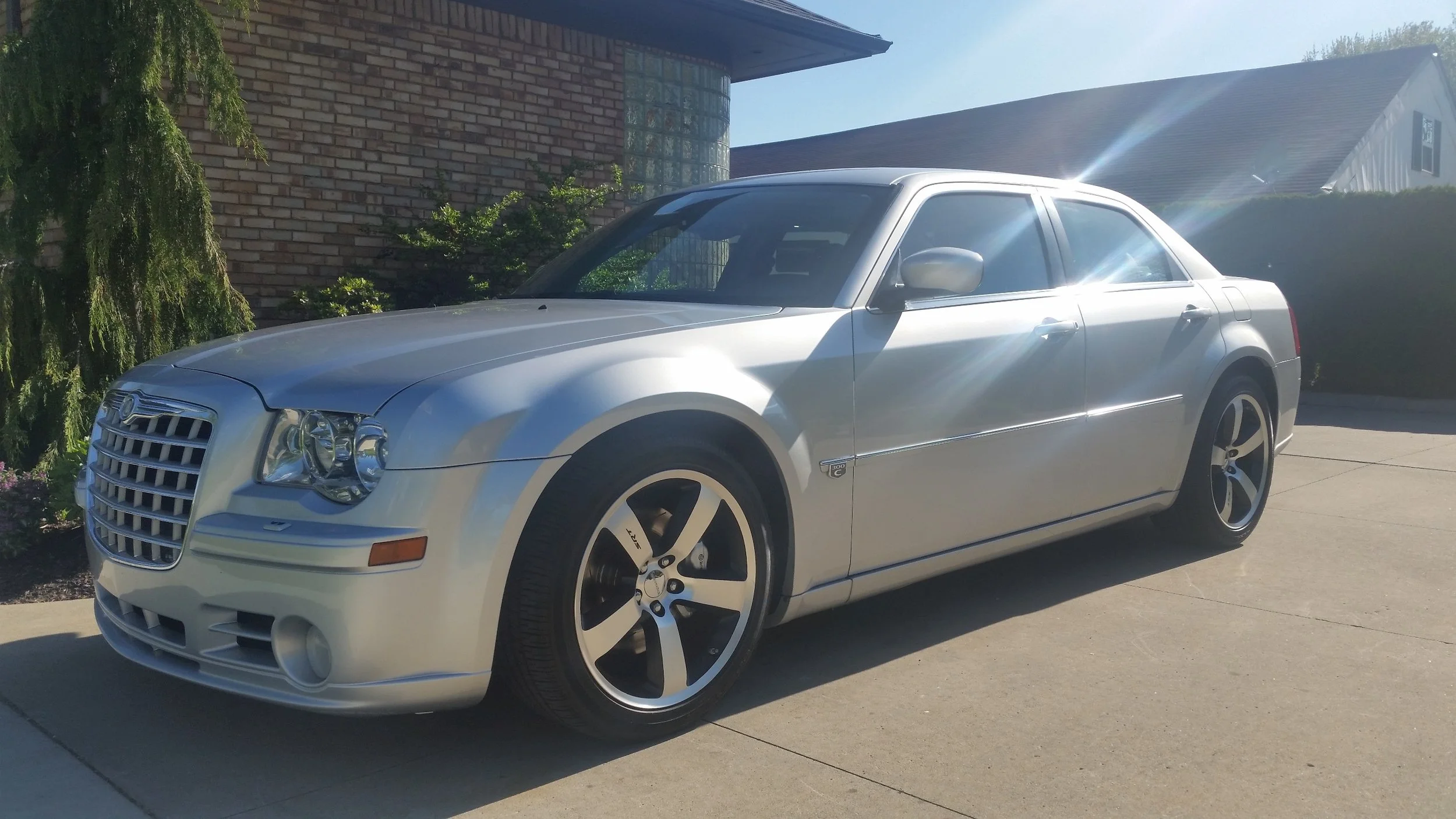 Silver sedan vehicle parked on driveway with house and trees in background, bright sunlight, and lens flare.