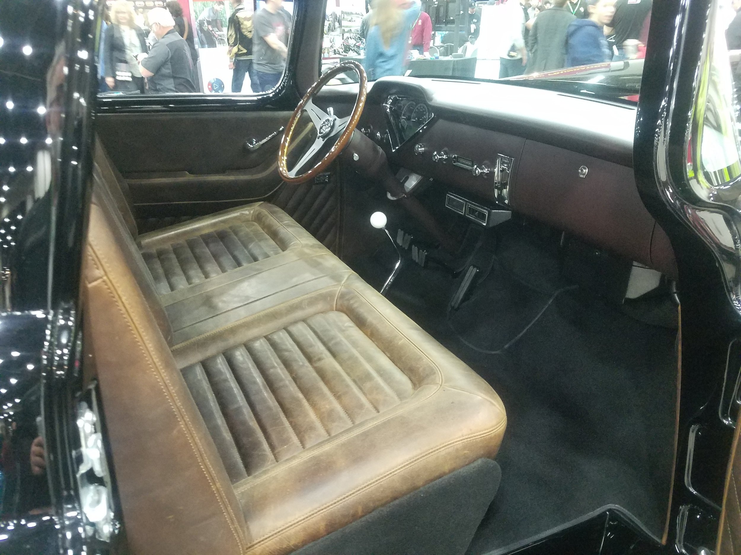 Interior view of a vintage car showing a brown bench seat, a dashboard with gauges, and a steering wheel with a wooden rim. The gear shifter is visible in the center.