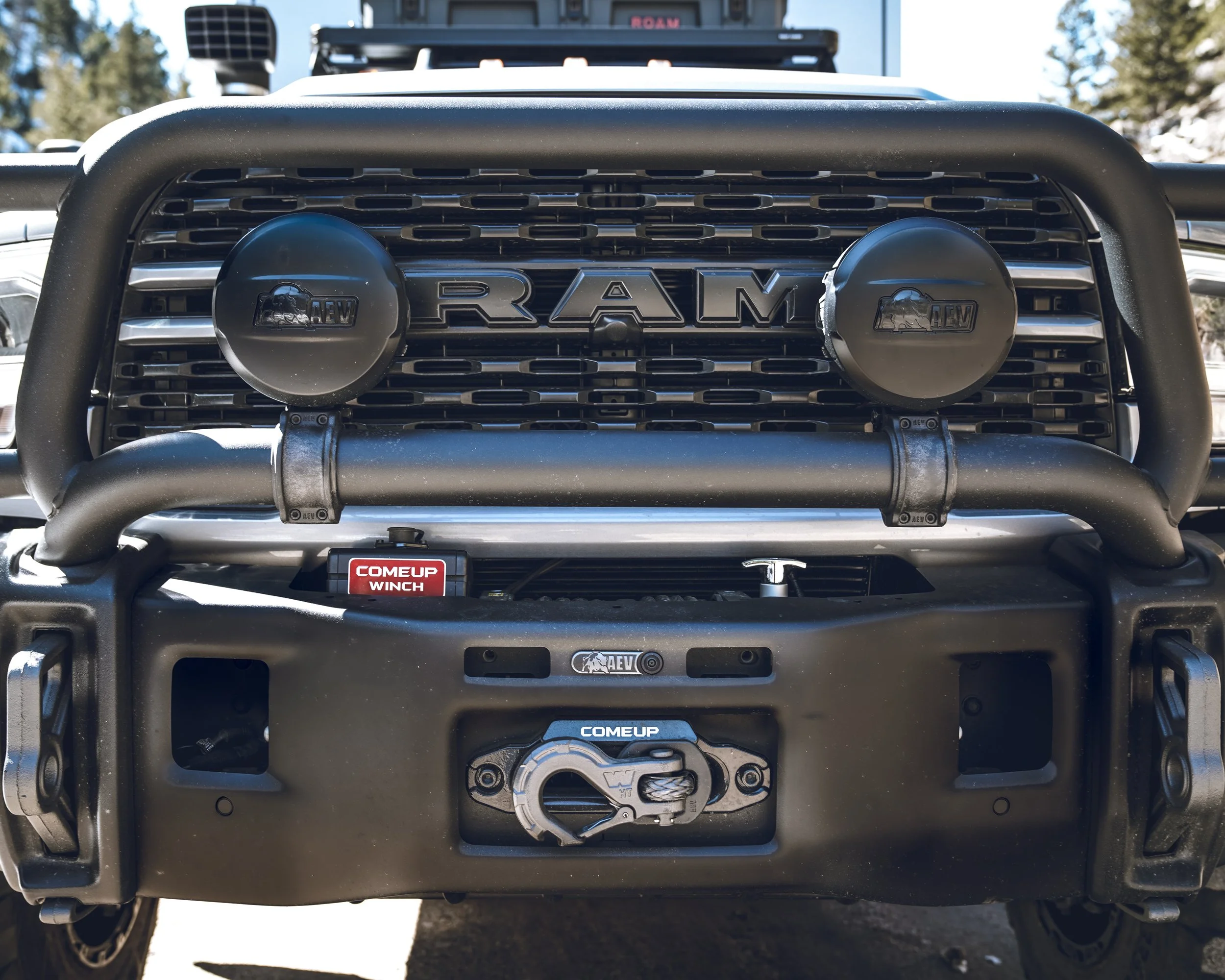 Close-up view of the front of a black RAM truck with off-road accessories, including a heavy-duty bumper, two large round auxiliary lights, a winch, and a grille with RAM lettering.