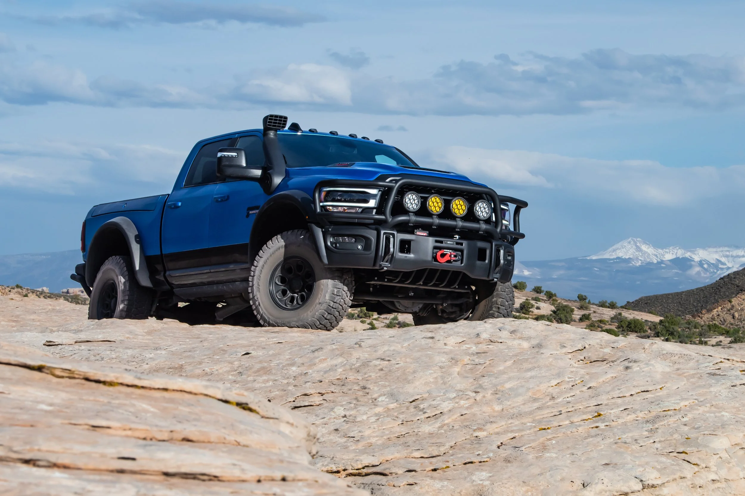 A blue pickup truck with off-road modifications parked on rocky terrain with mountains and partly cloudy sky in the background.