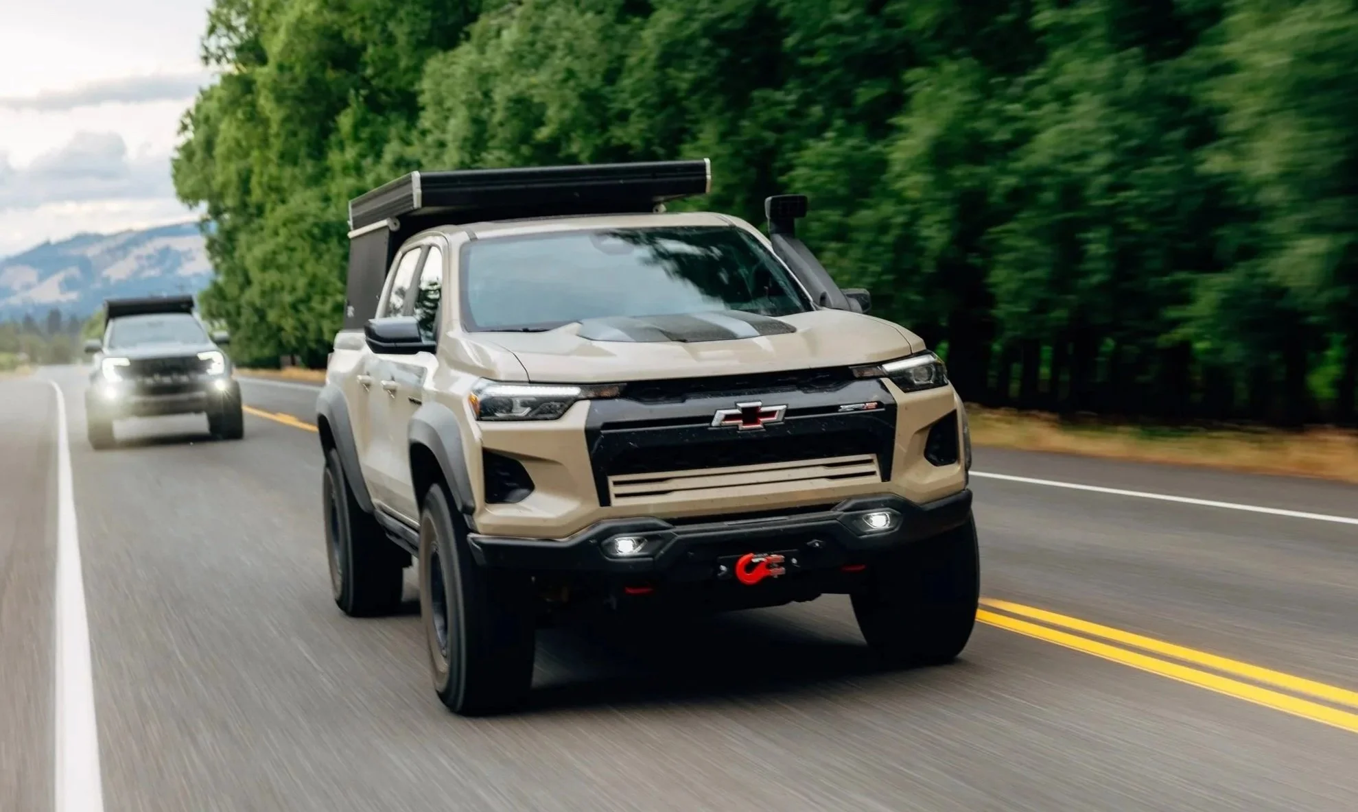 A beige Chevrolet SUV with off-road accessories including a roof rack and front bumper winch, driving on a two-lane road with a forested background and a mountainous landscape under a partly cloudy sky.