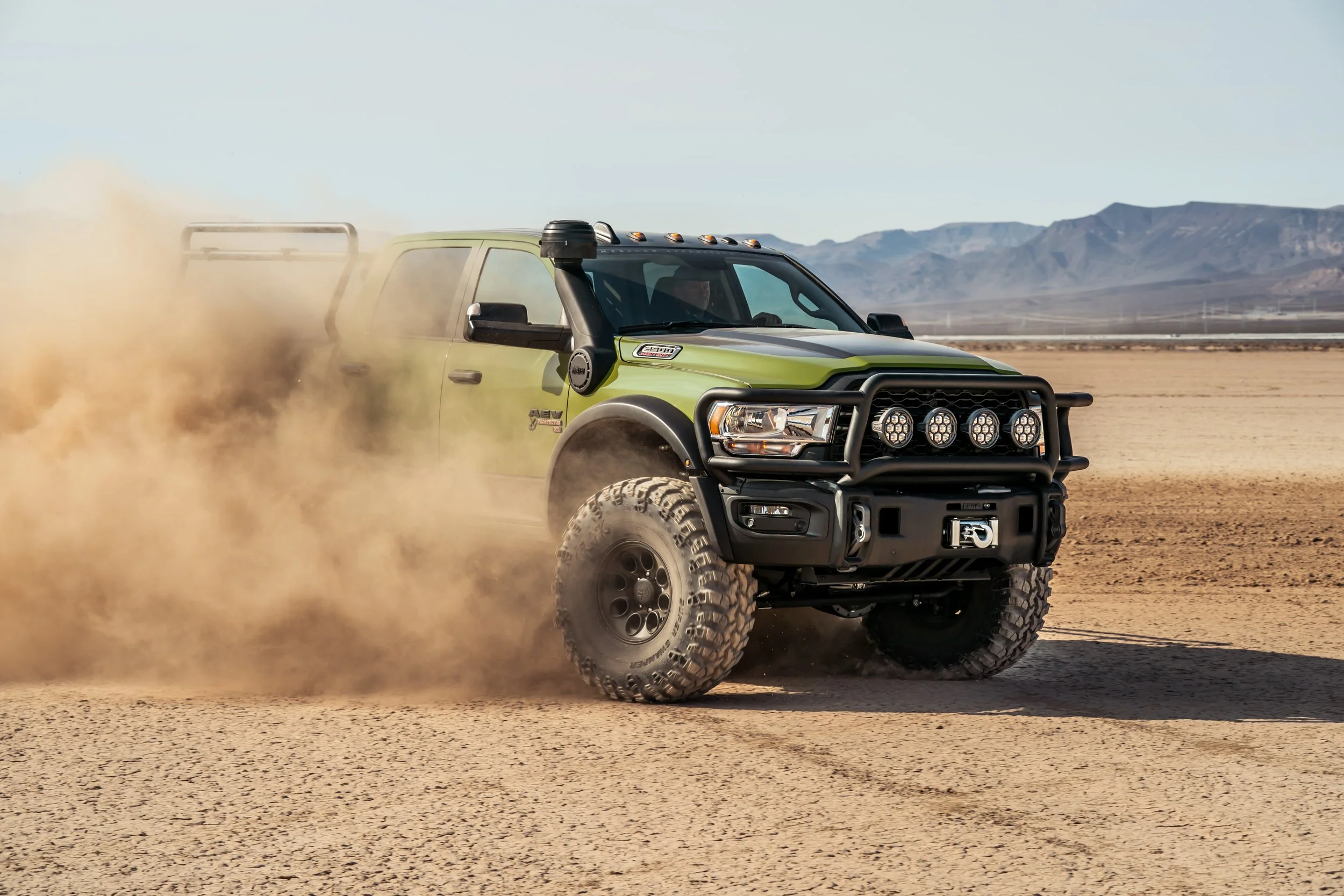 A green off-road truck driving through a desert, kicking up dust behind it.