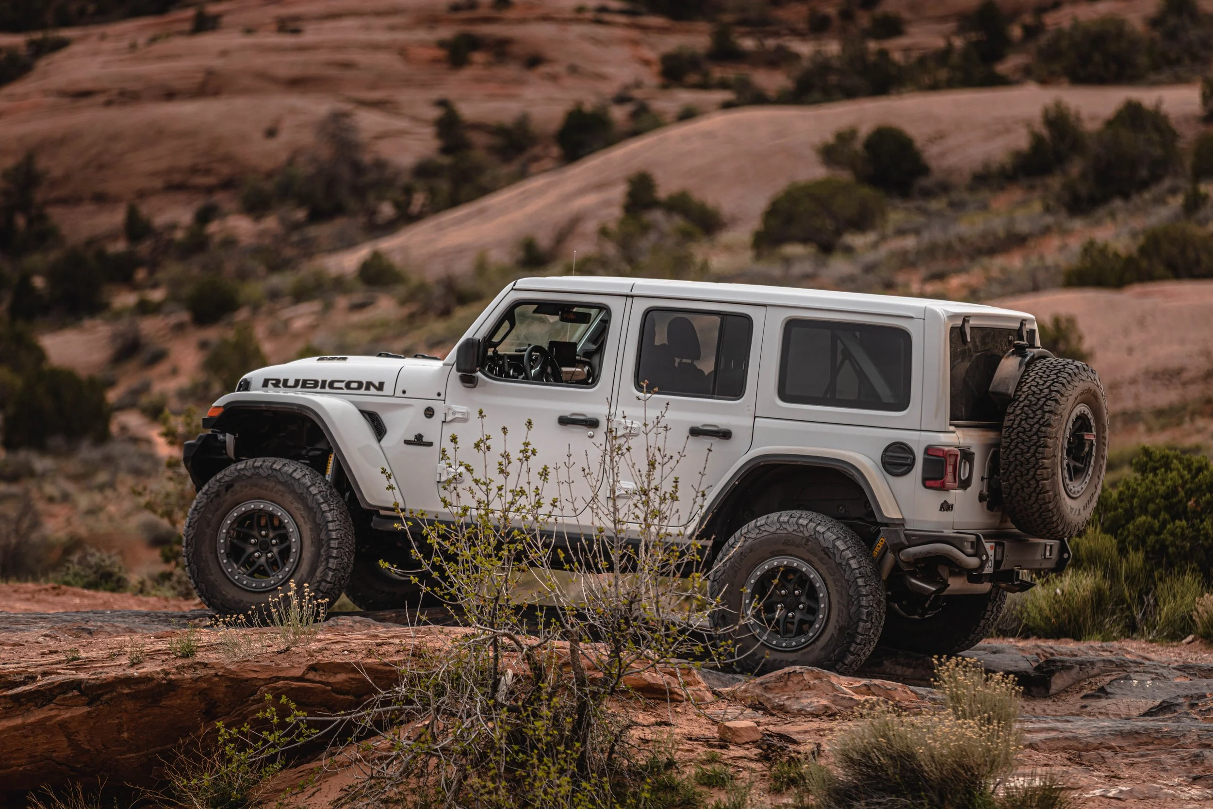 A white Jeep Rubicon 4x4 off-road vehicle on rocky terrain in a desert landscape with sparse bushes and hills in the background.