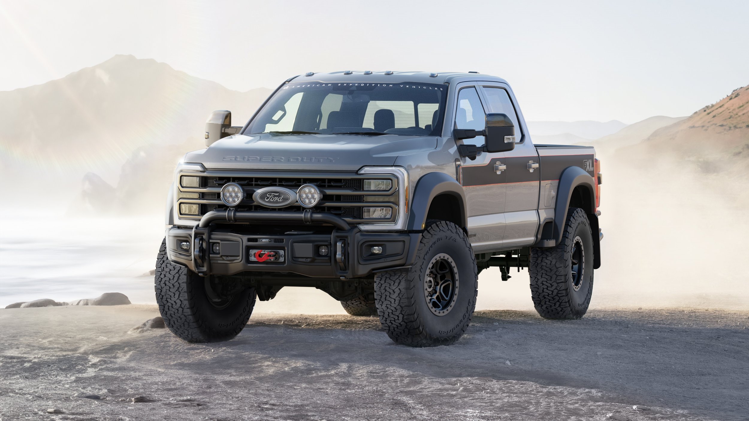 Gray Ford Super Duty pickup truck with off-road tires, front bull bar, and off-road lights, parked on a dirt landscape with mountains and dust in the background.