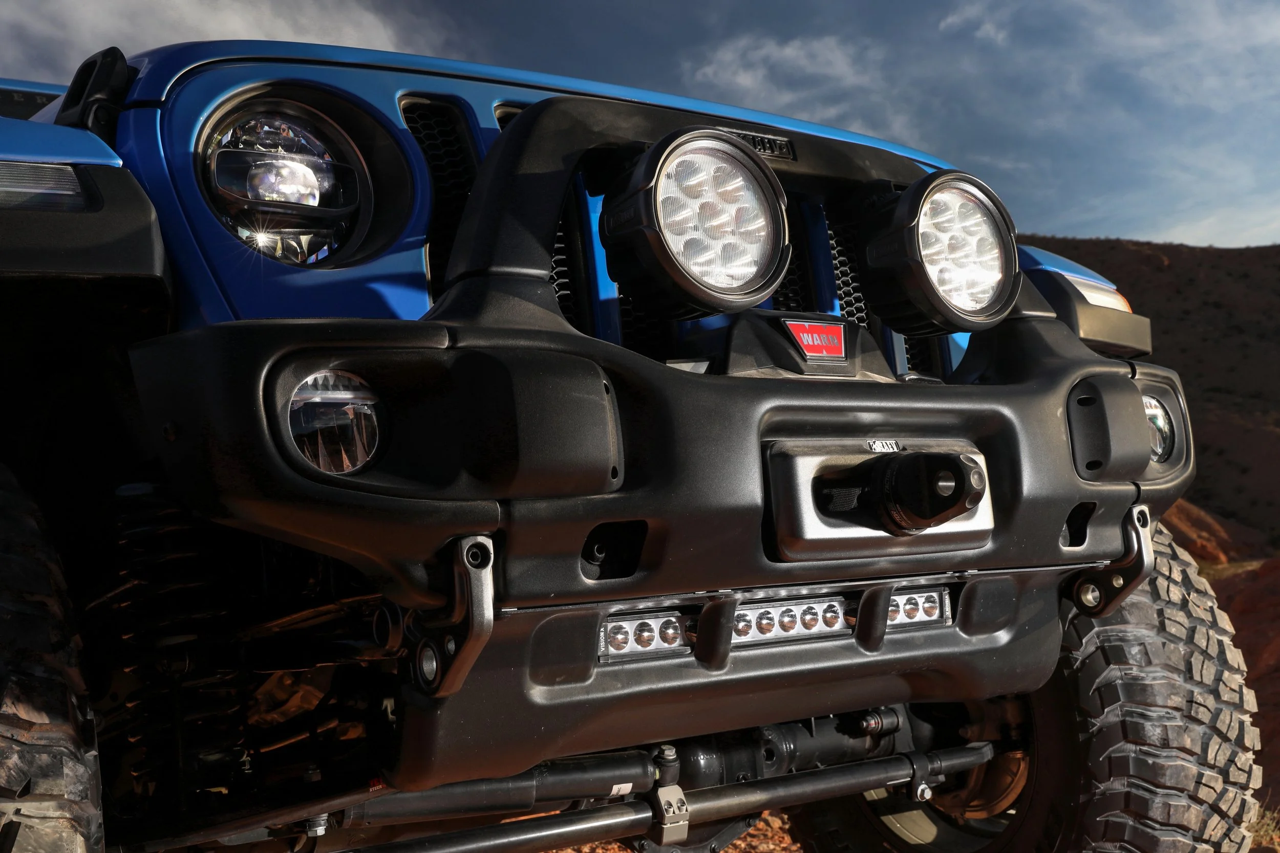 Close-up of the front of a blue off-road vehicle with large tires, multiple headlights, and an off-road bumper, set against a mountainous landscape and cloudy sky.
