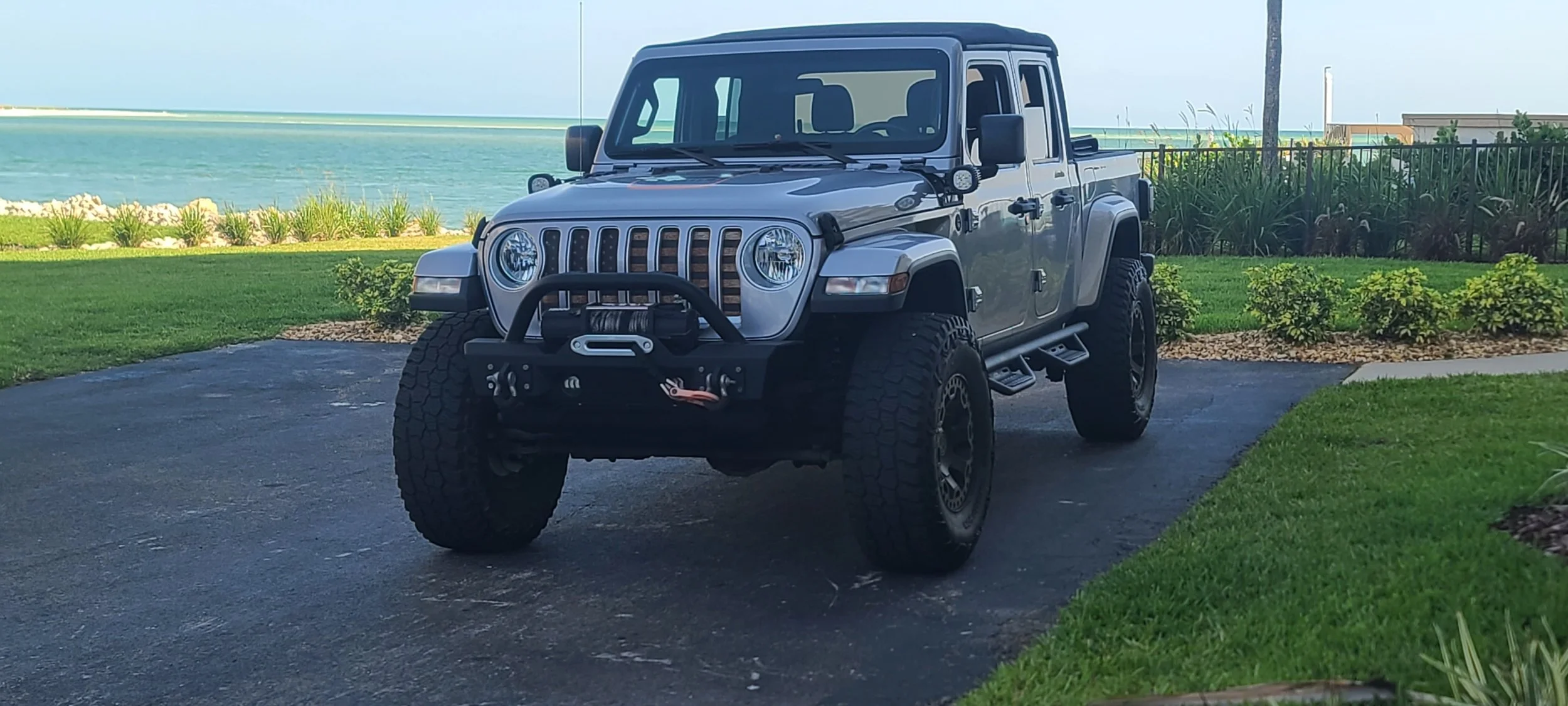 A silver Jeep Gladiator truck parked on a driveway near a grassy area with a waterfront view in the background.