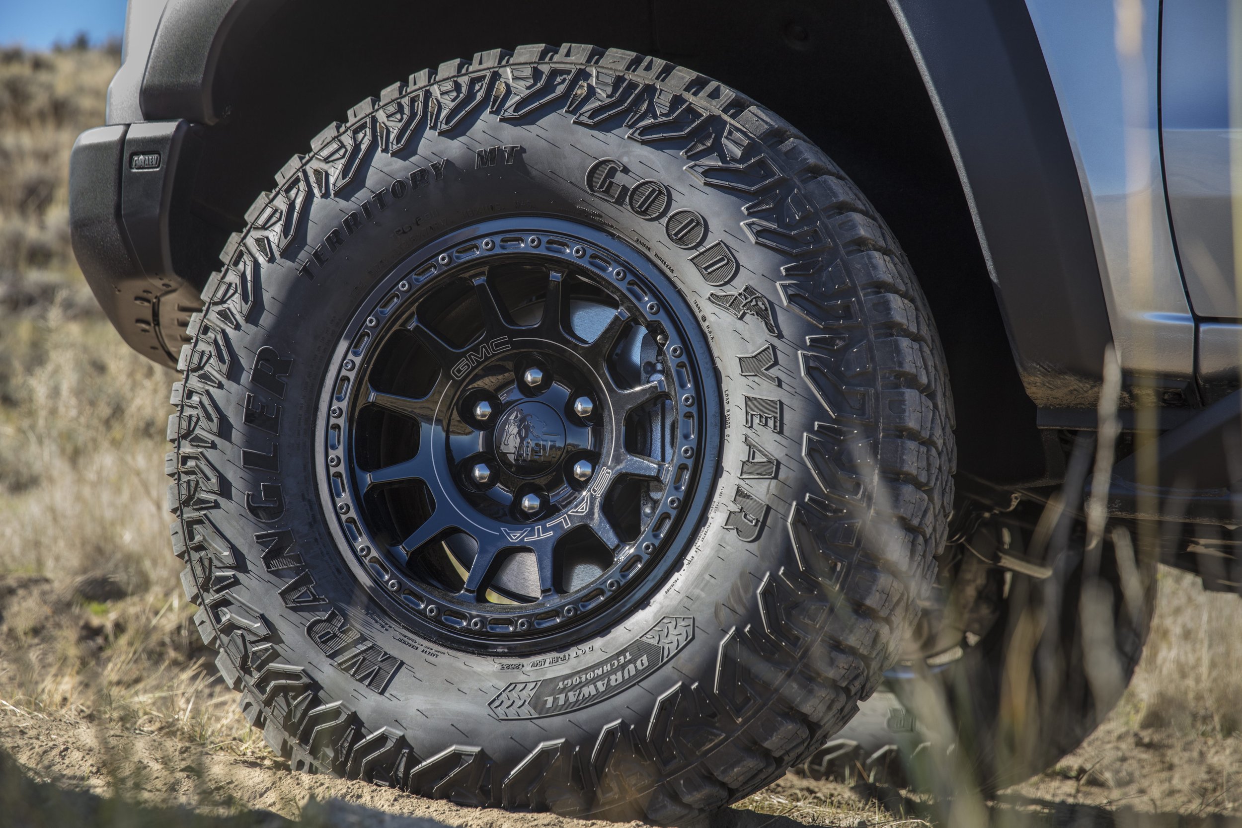 Close-up of a rugged off-road tire on a vehicle, with the brand name 'Goodyear' and model 'Wrangler All-Terrain Adventure' visible on the tire, along with the black rim and part of the vehicle's body.