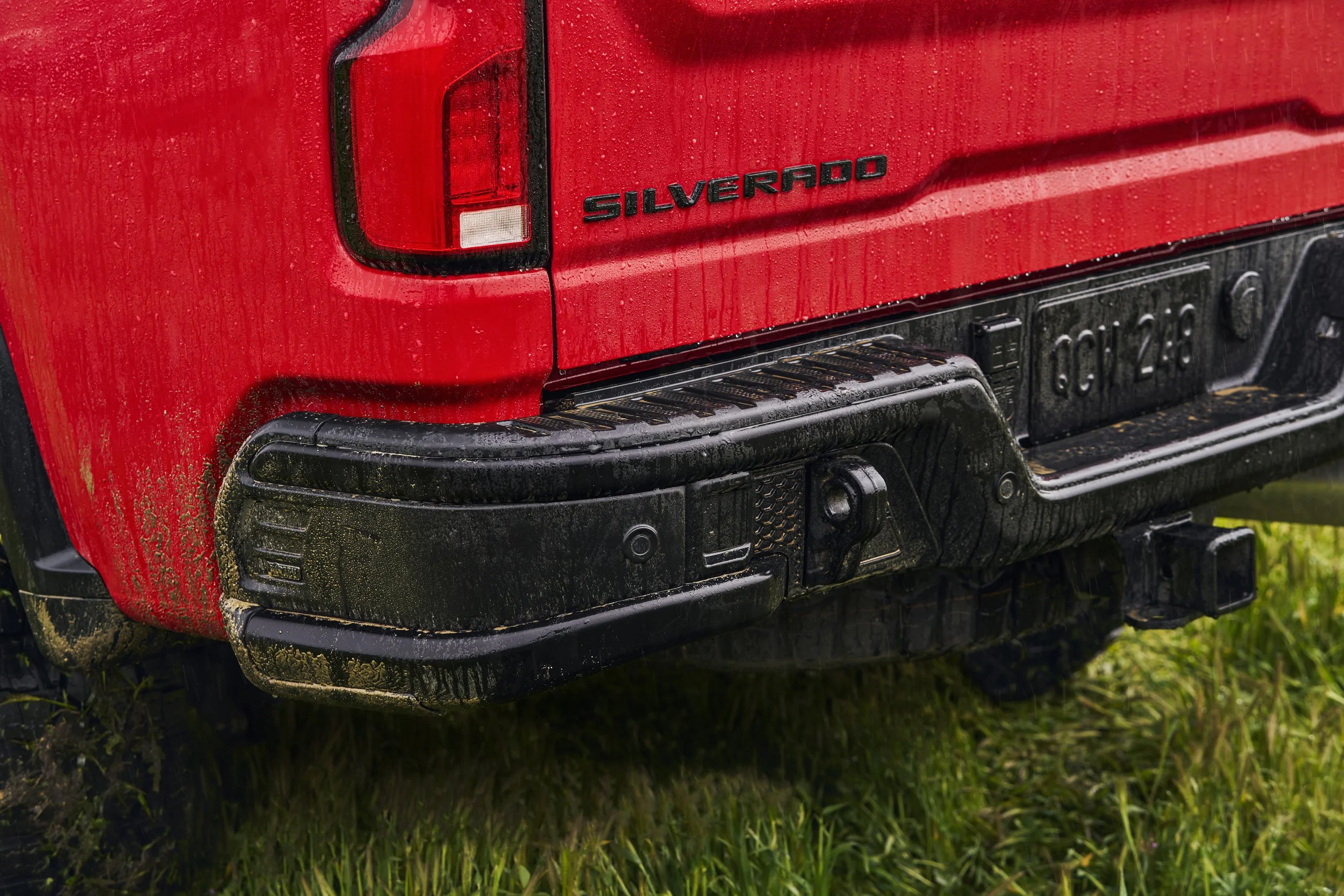 Close-up of the back of a red Chevrolet Silverado truck showing the tail light, black bumper, and Silverado badge, with dirt on the bumper and grass underneath.