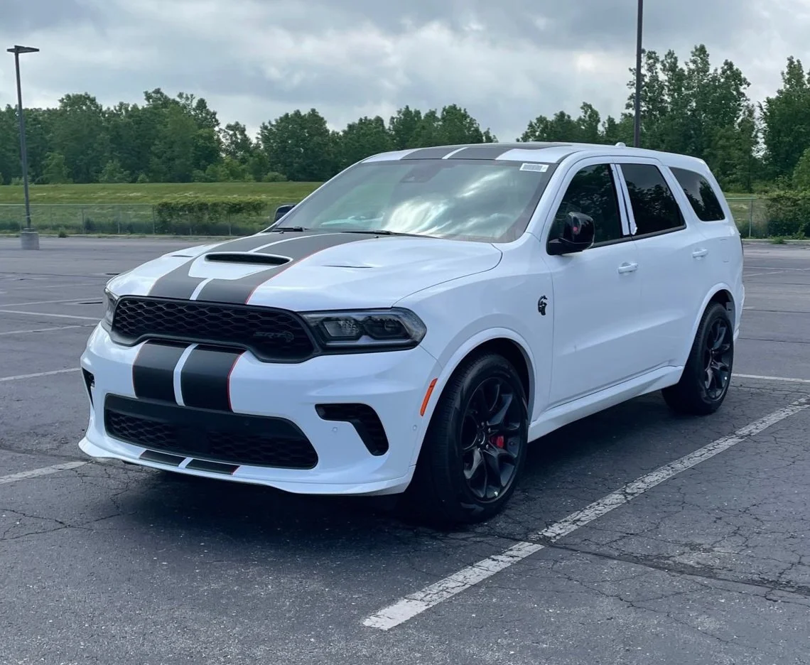 A white SUV with black racing stripes parked in an empty parking lot under cloudy skies, surrounded by green trees in the background.