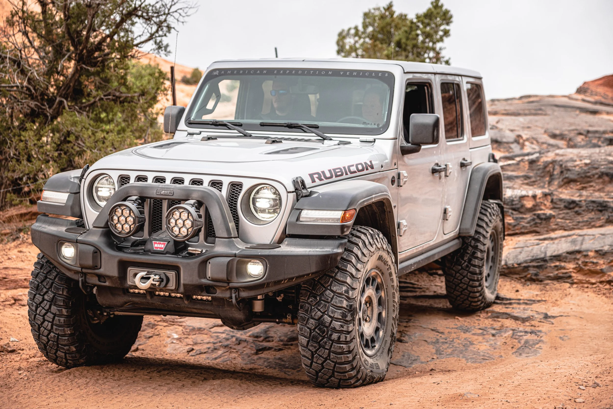 A silver Jeep Rubicon off-road vehicle parked on a dirt trail in a desert landscape with rocks and sparse vegetation.