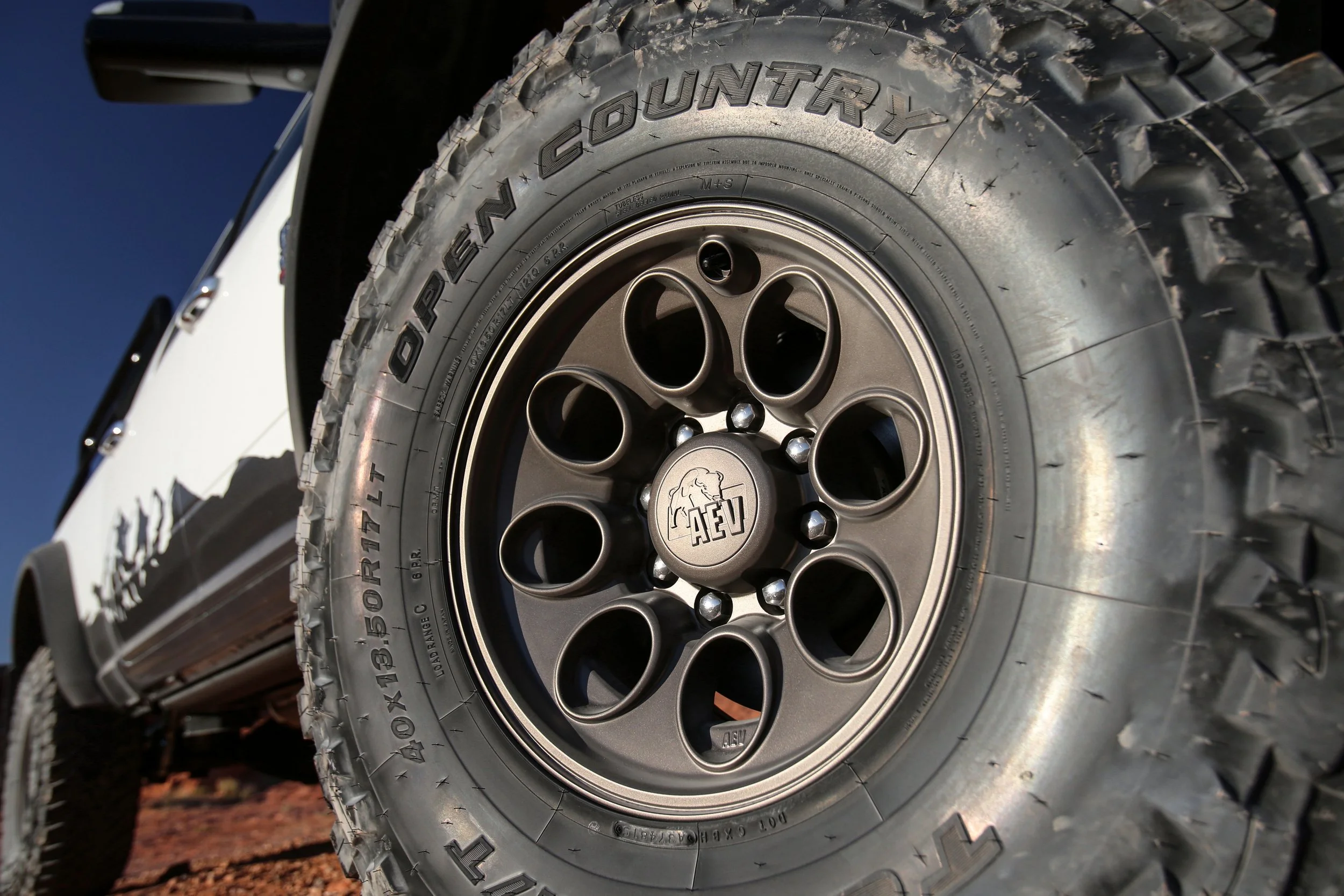 Close-up of a tire and wheel on a white off-road vehicle with an AE V logo on the center cap, set against a clear sky and rugged terrain.
