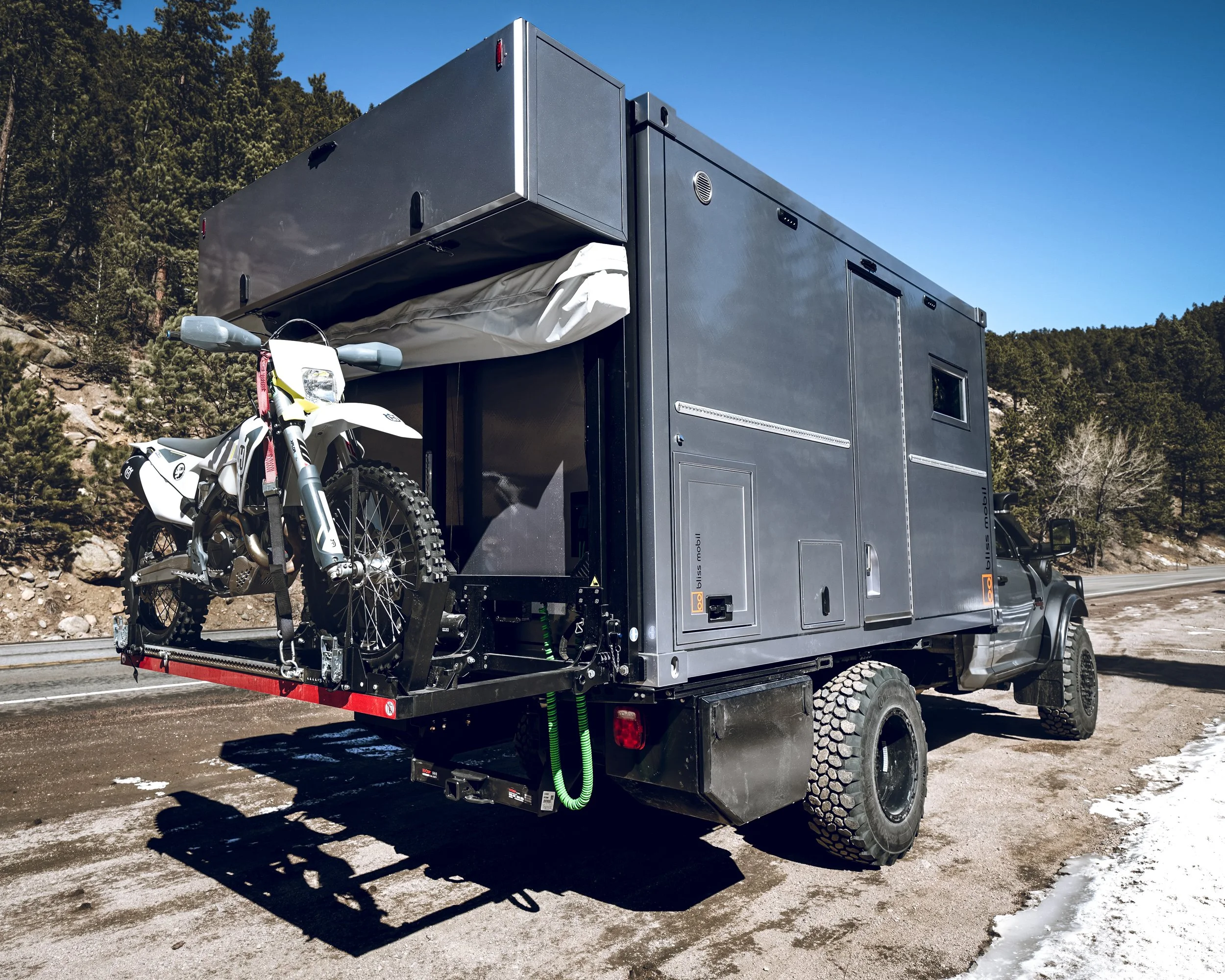A large black off-road truck with a Moto Cross dirt bike mounted on the rear cargo platform, parked on a roadside with trees and mountains in the background.
