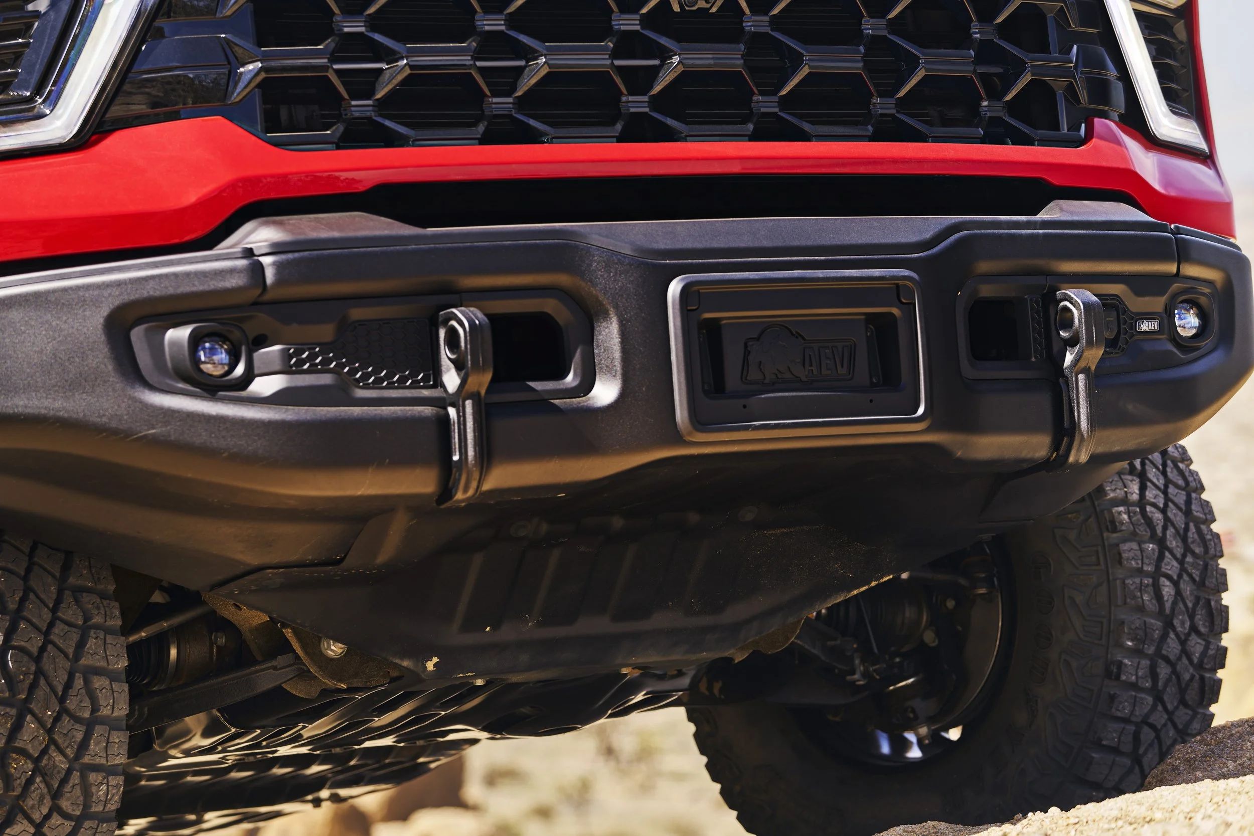 Close-up of the front bumper of a red off-road vehicle with a black grille, winch hooks, and rugged tires.