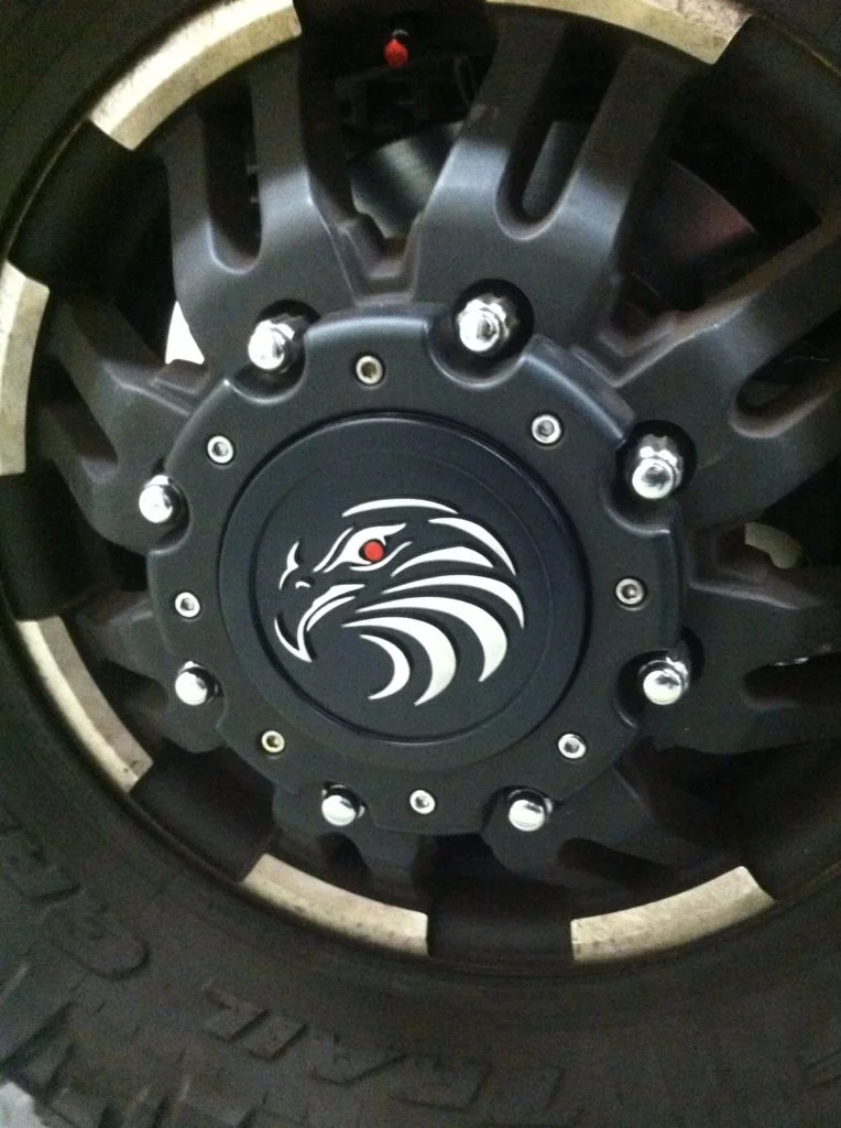 Close-up of a black car wheel with a hubcap featuring an eagle head logo and bolt cover nuts.