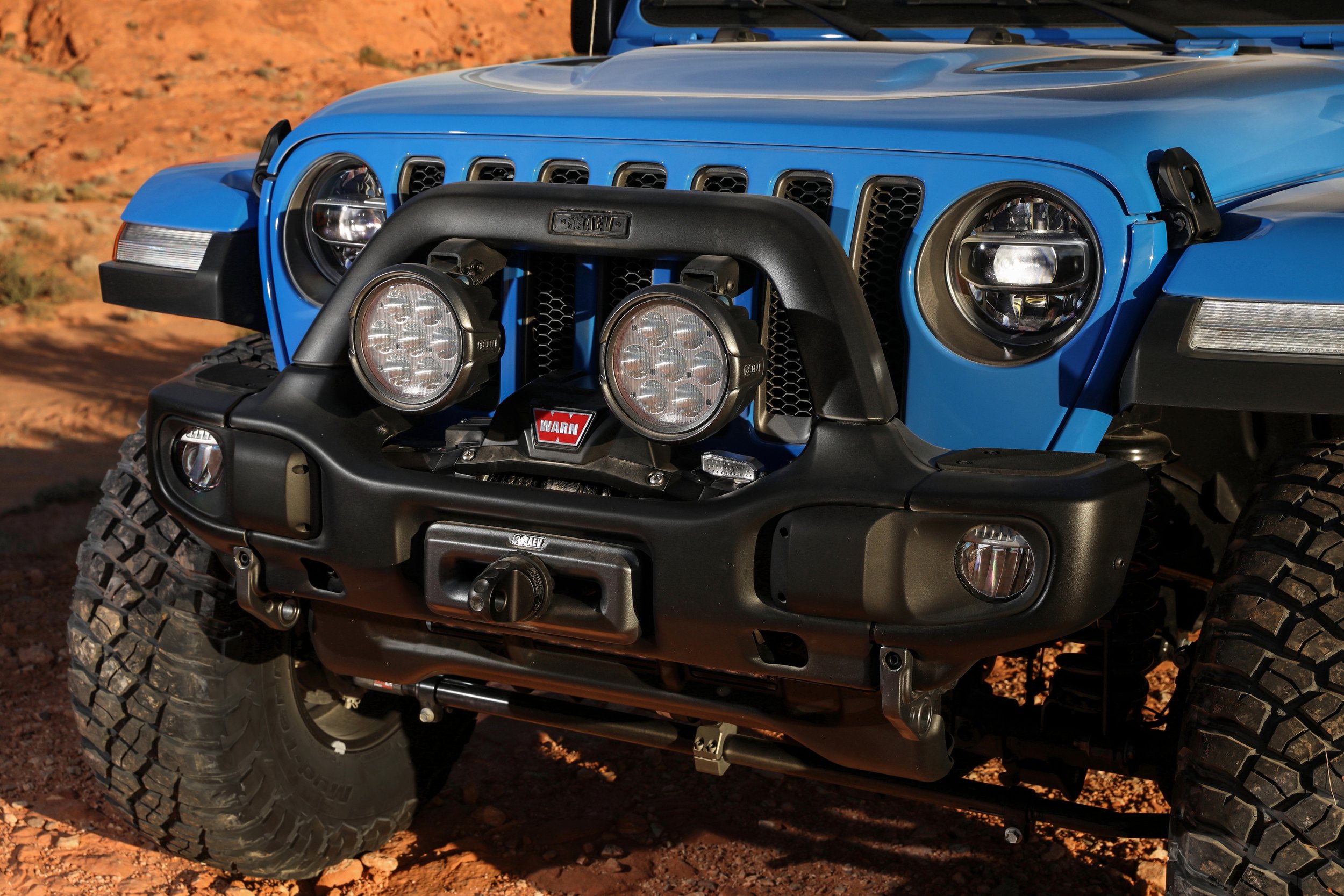Front view of a blue off-road Jeep with a black bumper, large tires, and multiple round auxiliary lights mounted on the bumper and grille, parked on dirt terrain.