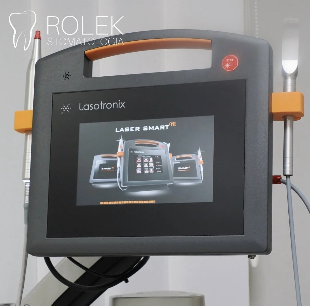 A Lasotronix Laser Smart medical device with a digital display showing the device's interface. The device has a handle on top, orange accents, and is connected to a medical tool on each side with orange holders. The background is plain, likely in a medical or clinical setting.