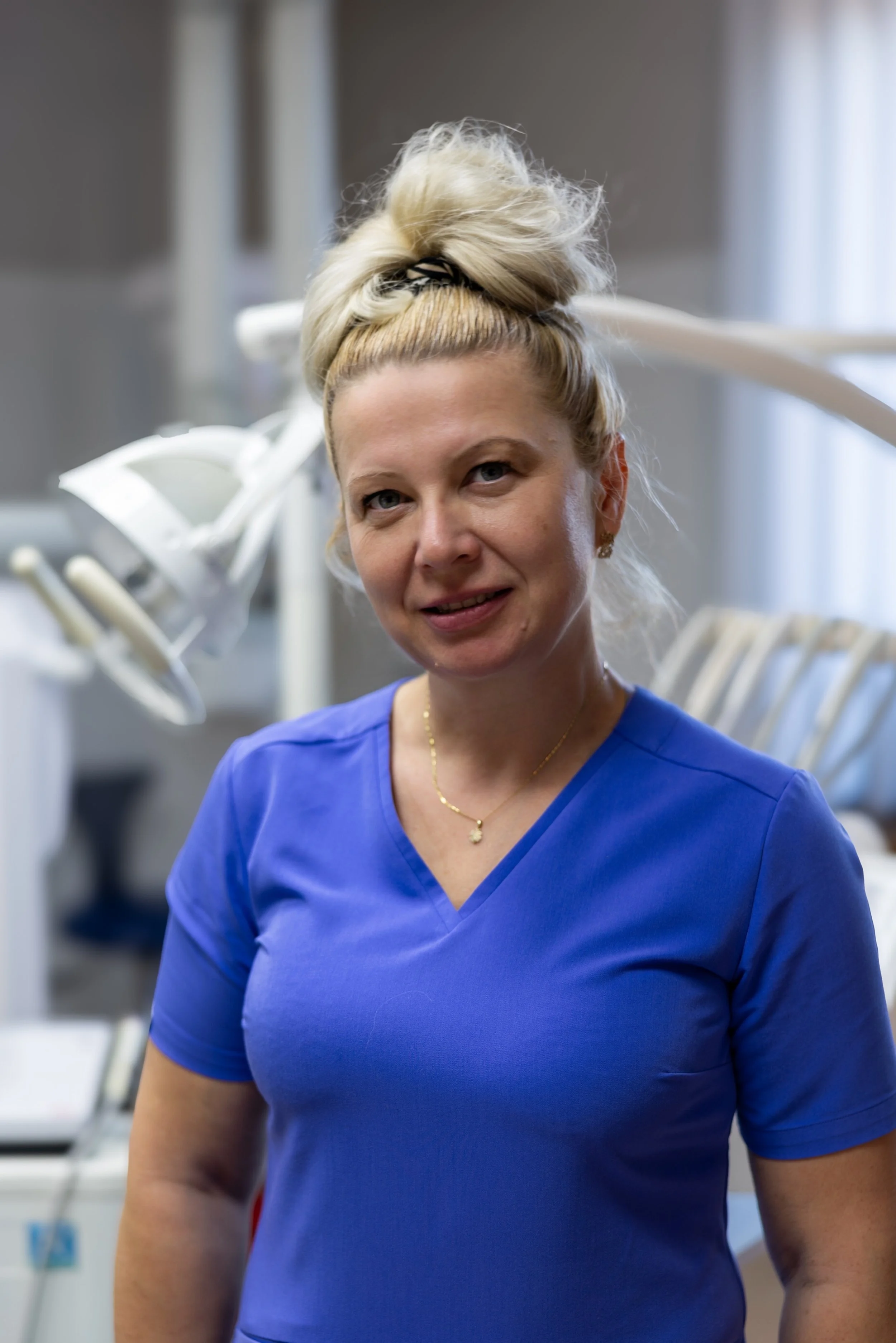 A woman dressed in blue medical scrubs stands in a clinical or dental setting with dental equipment in the background.