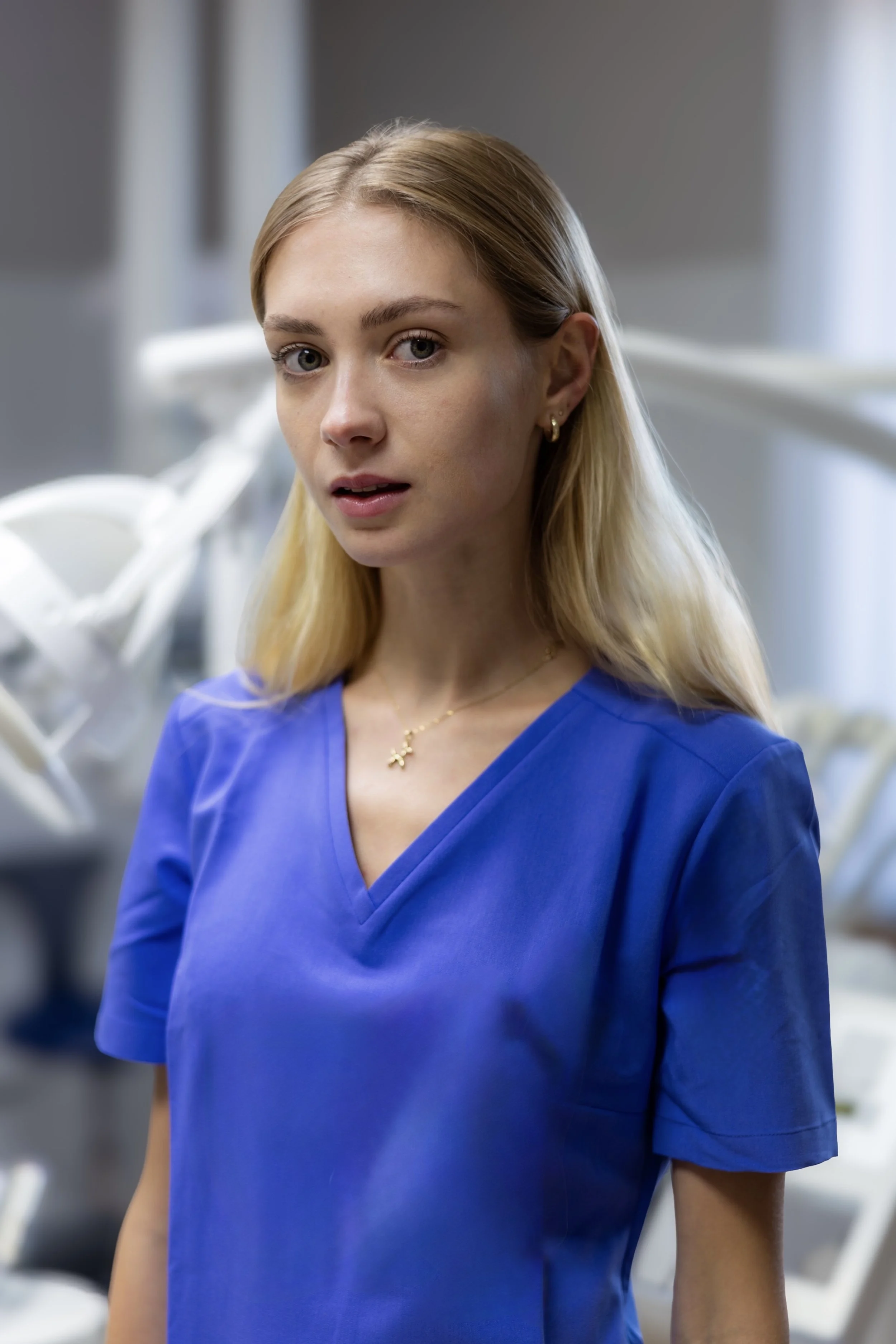 A young woman in blue medical scrubs standing in a clinical setting, looking at the camera with a neutral expression.