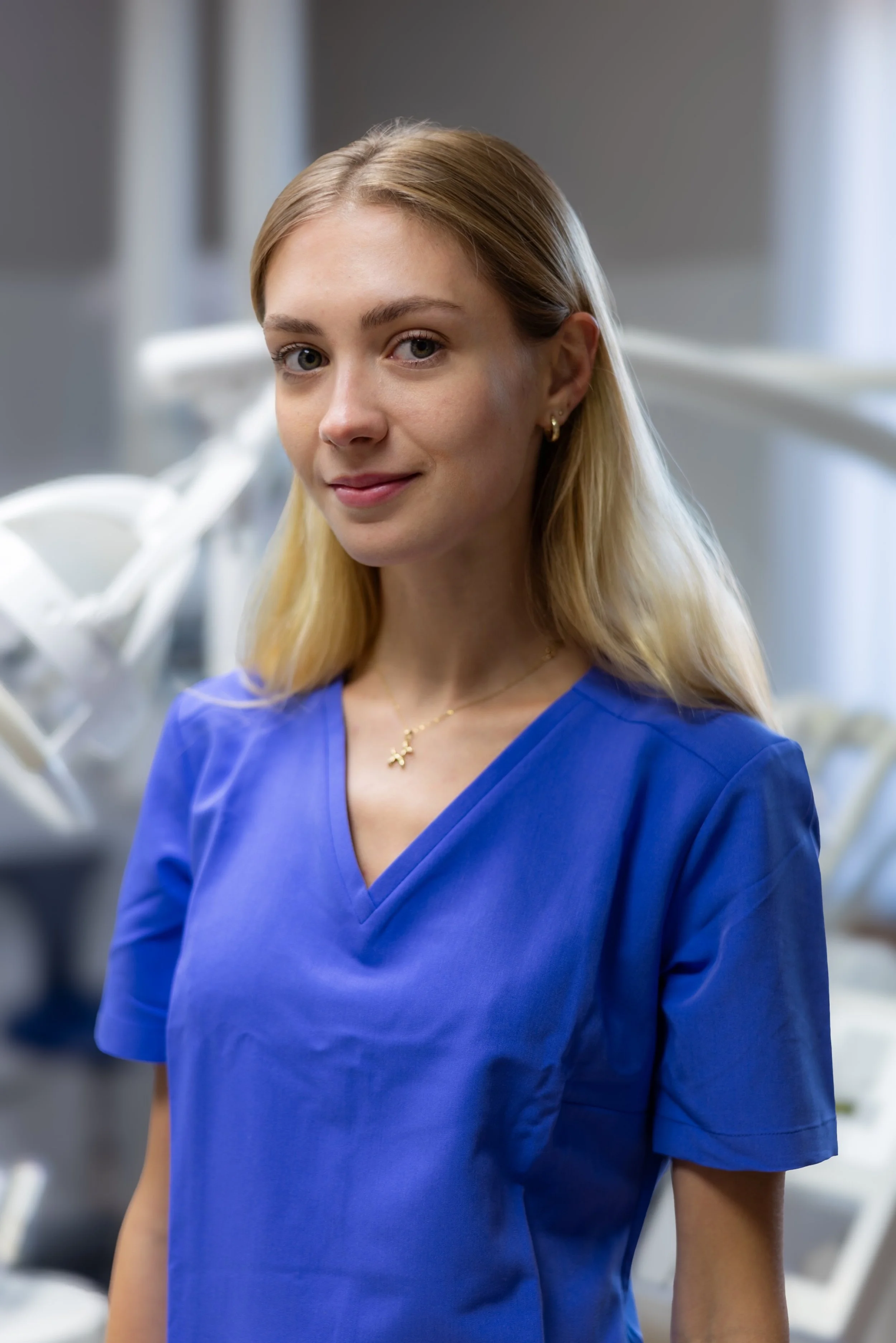 A young woman with blonde hair, wearing blue scrubs, standing in a medical or laboratory setting with equipment in the background.