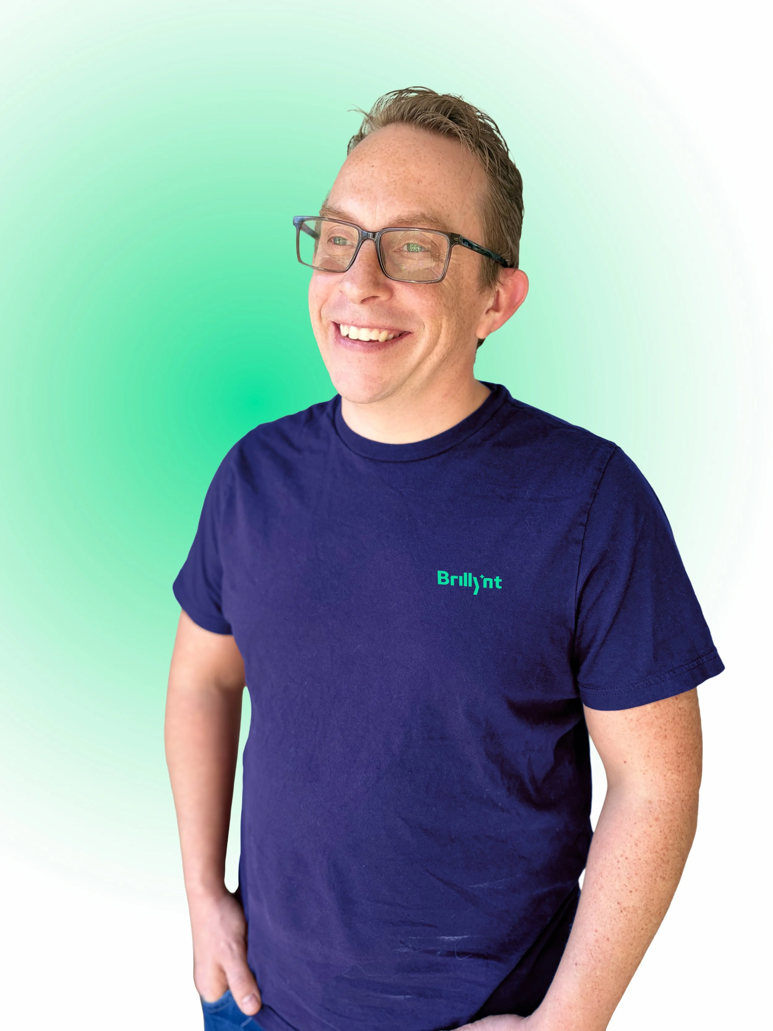 Smiling man with glasses wearing a navy blue t-shirt with the Brillynt logo, standing against a gradient green background.