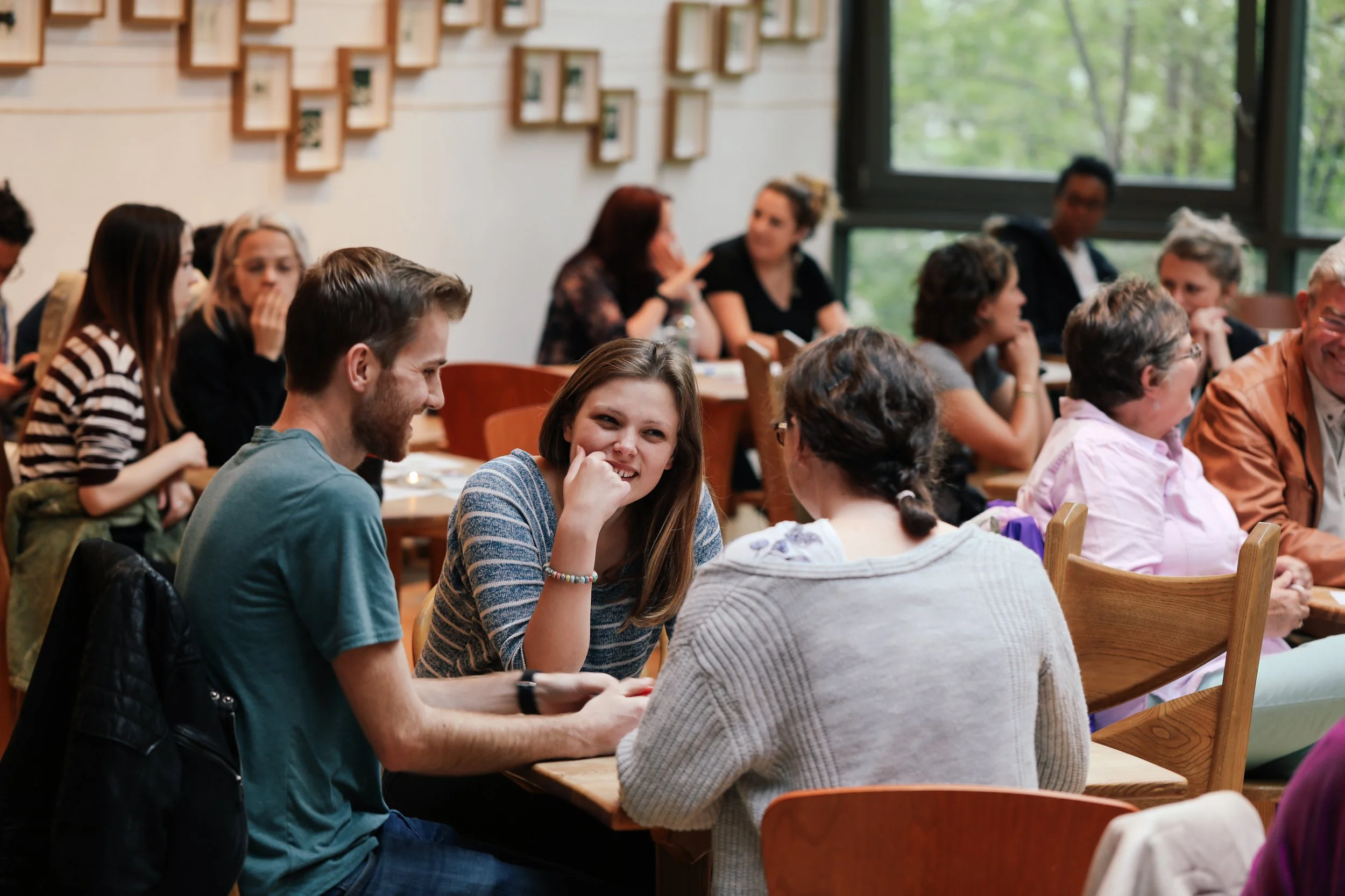 A group of people sitting at tables in a room with large windows, engaging in conversation.