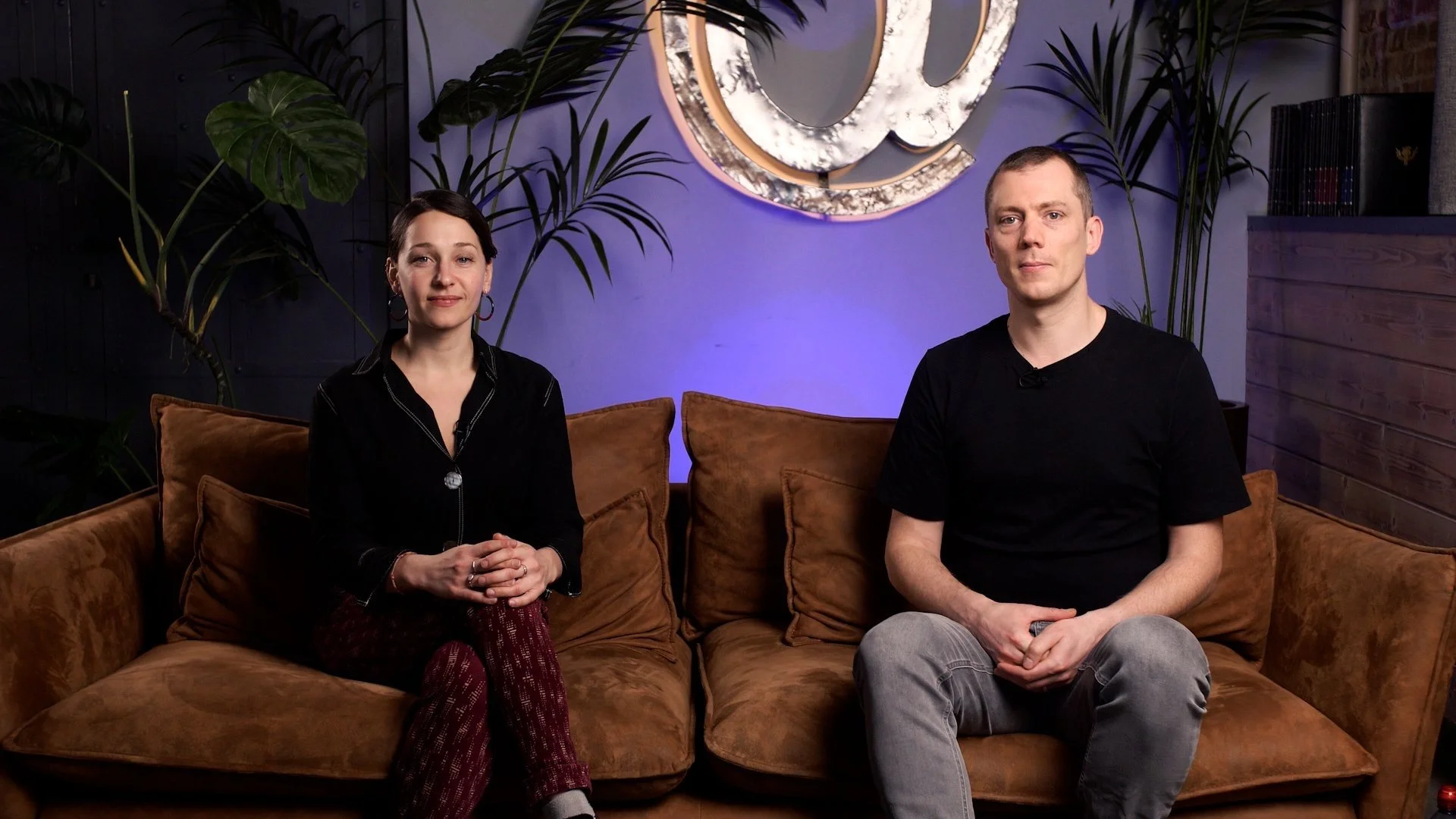 A woman and a man sitting on a brown sofa in a room with blue lighting and plants, with a decorative round wall art piece behind them.