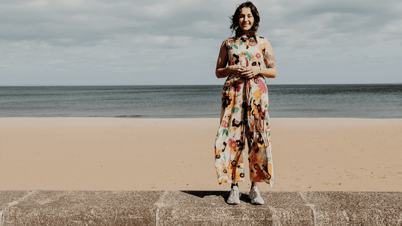 Lily Asch standing on a stone promenade at the beach, wearing a colorful floral jumpsuit and sneakers, smiling with hands clasped, with ocean and cloudy sky in the background.