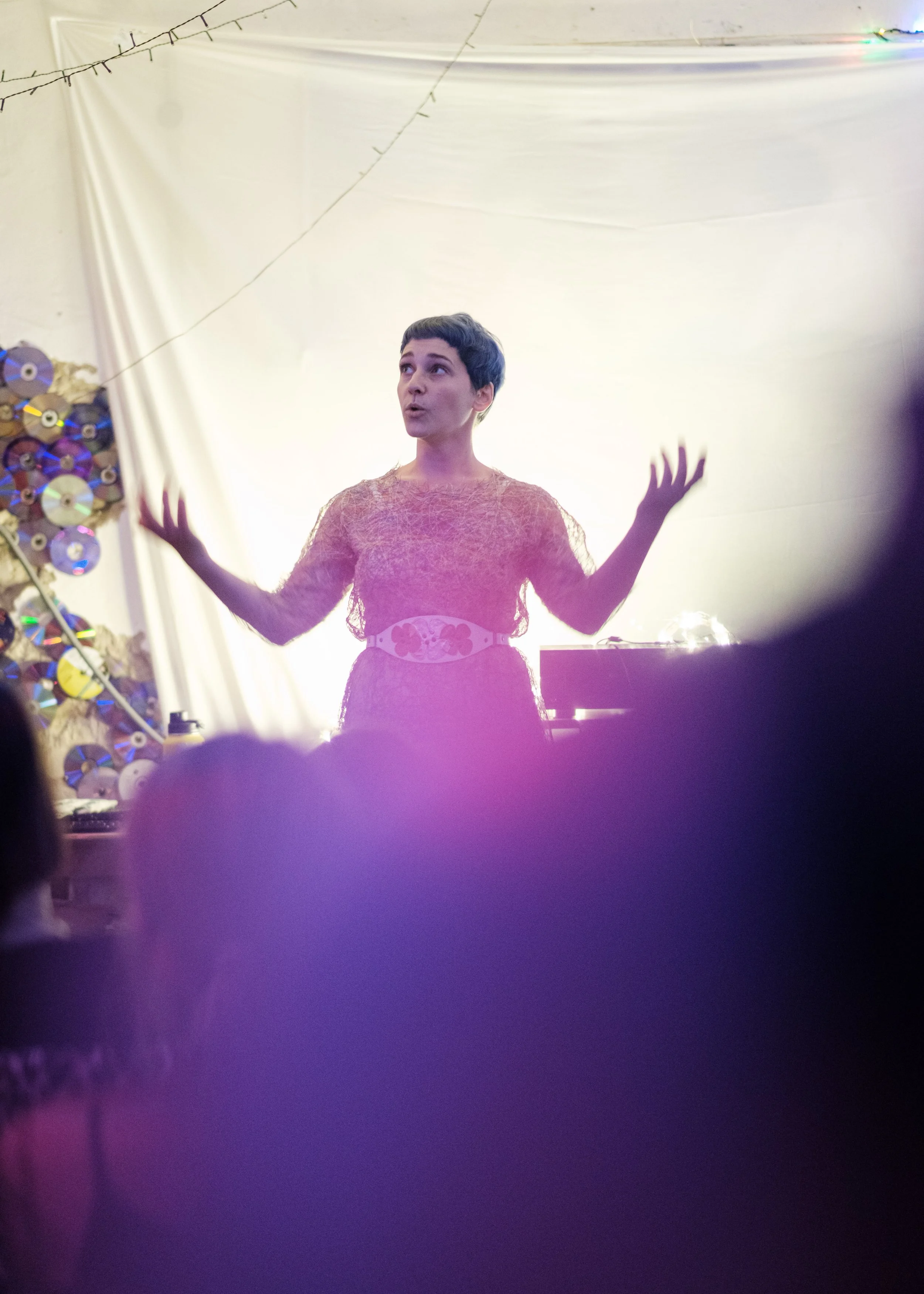 Person performing on stage at an event, behind a white backdrop with decorations made of CDs, with purple and yellow lighting.
