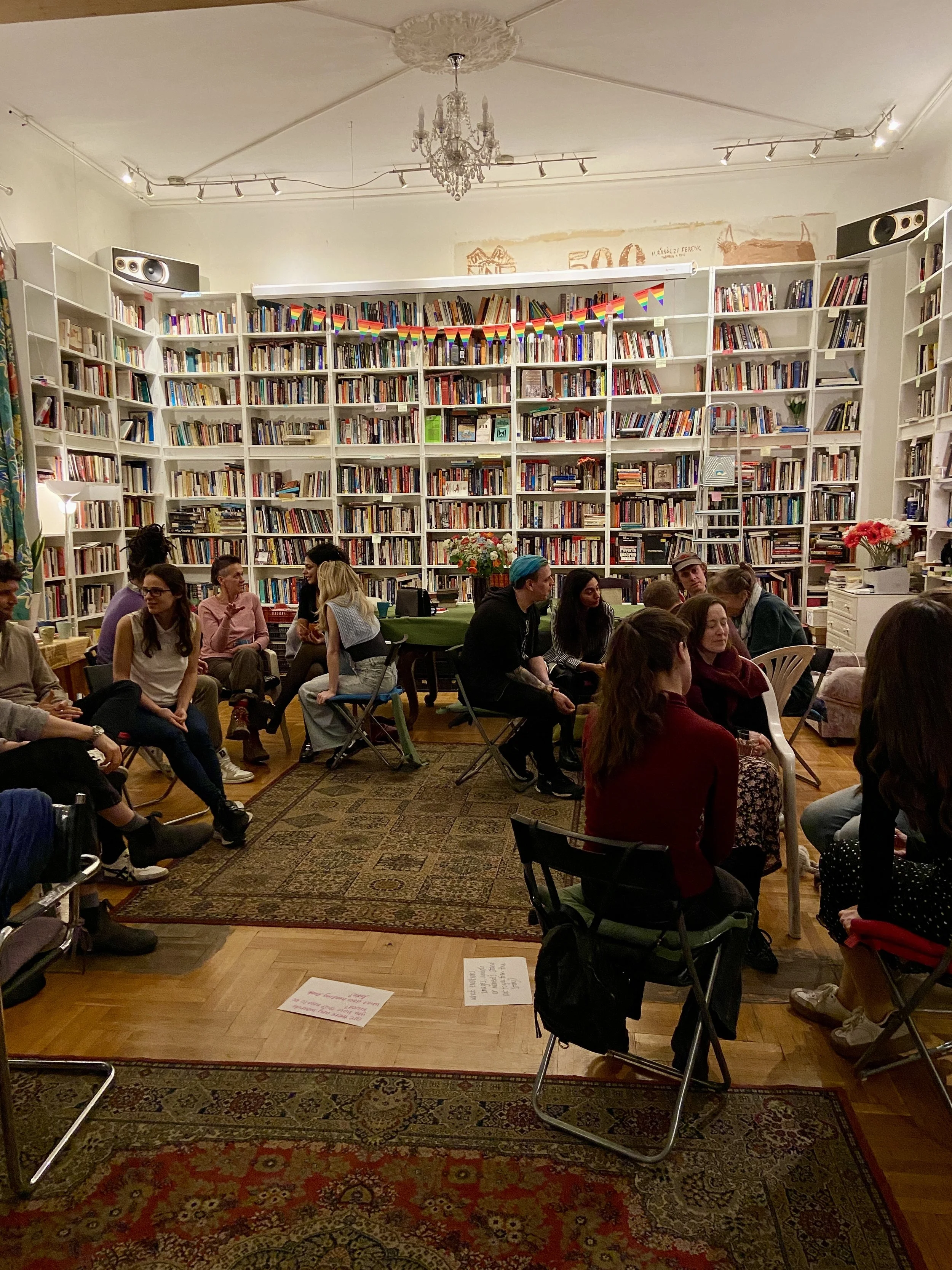 People sitting in a circle in a cozy room with tall bookshelves filled with books, a chandelier hanging from the ceiling, and a small table with a floral arrangement.