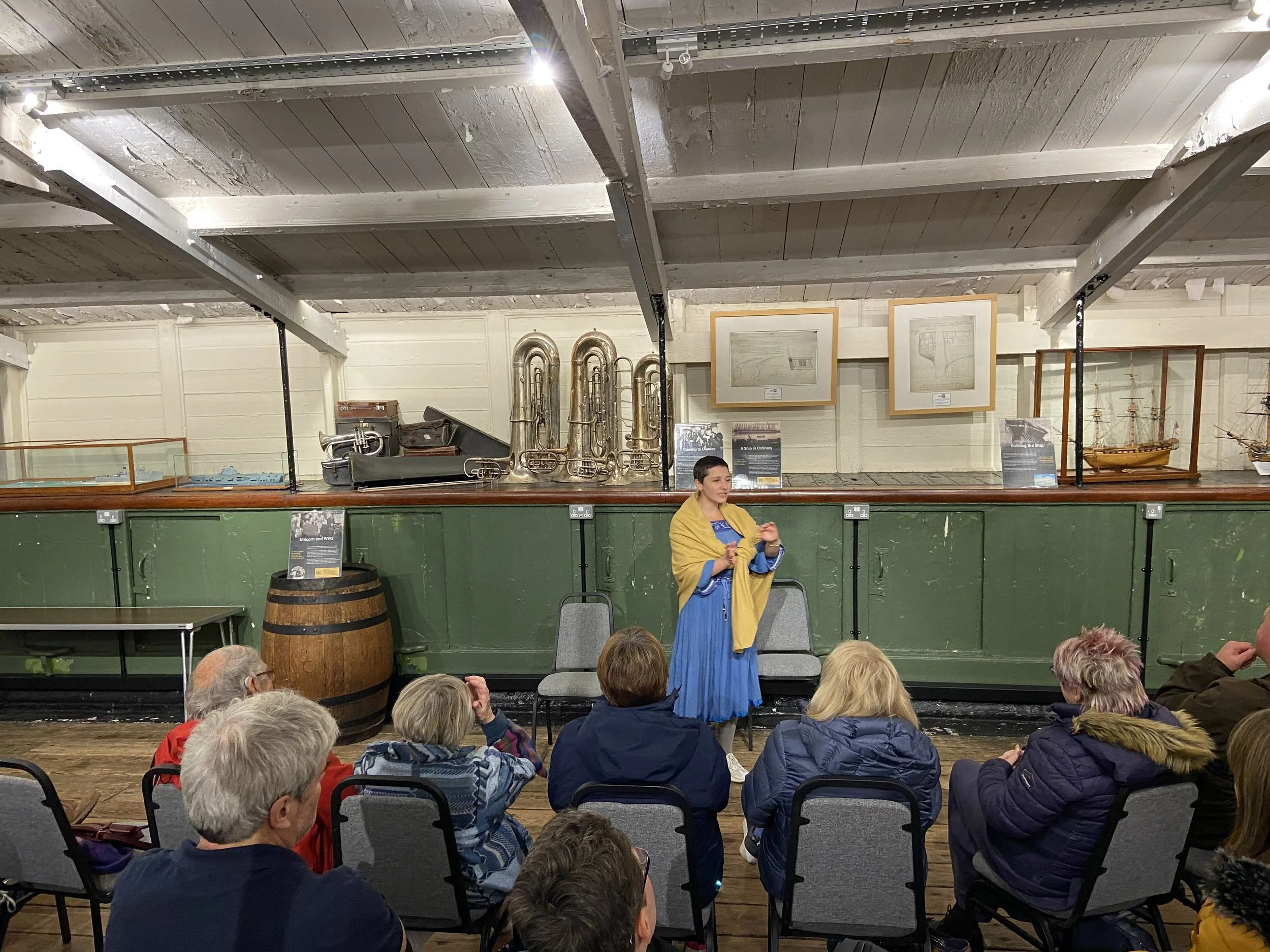 A woman in a blue dress and yellow shawl speaking to an audience seated in chairs inside a museum-like setting with display cases of model ships and historical artifacts on the wall behind her.