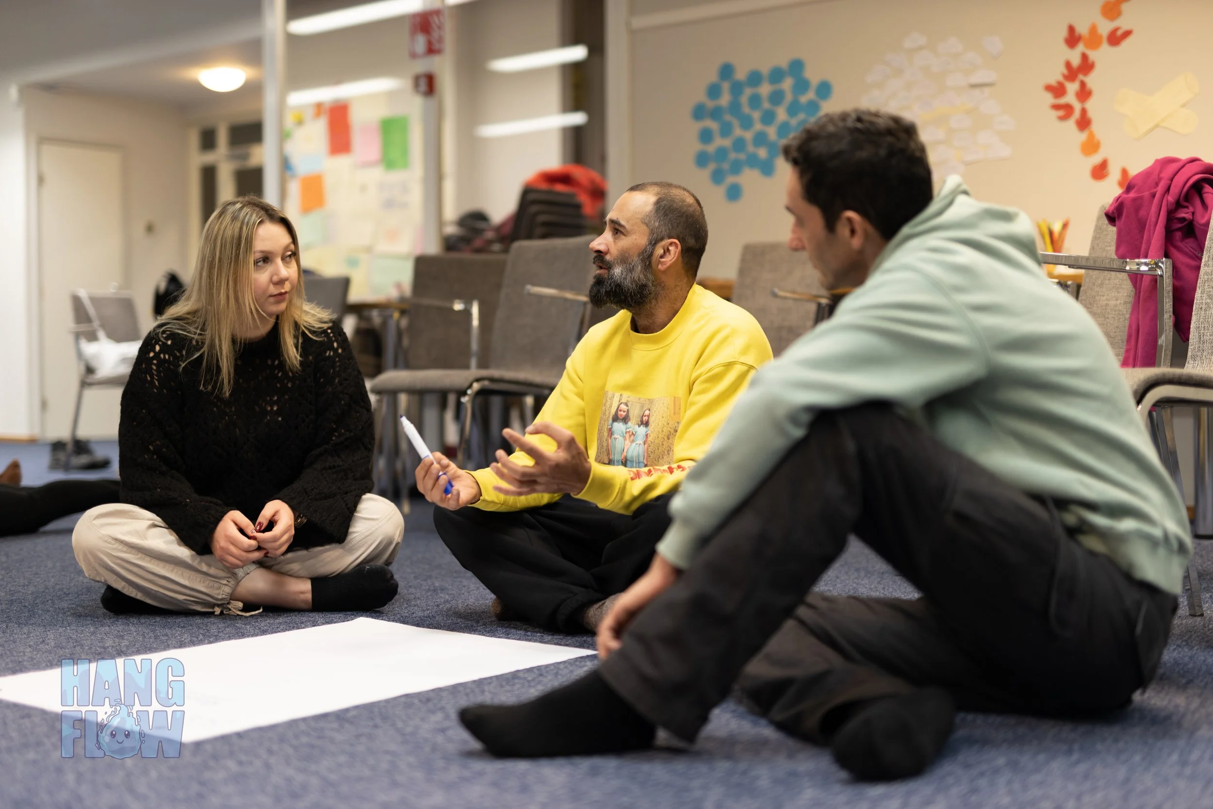 Three people sitting cross-legged on the carpet in a circle having a discussion in a room with chairs, backpacks, and colorful paper cutouts on the wall.