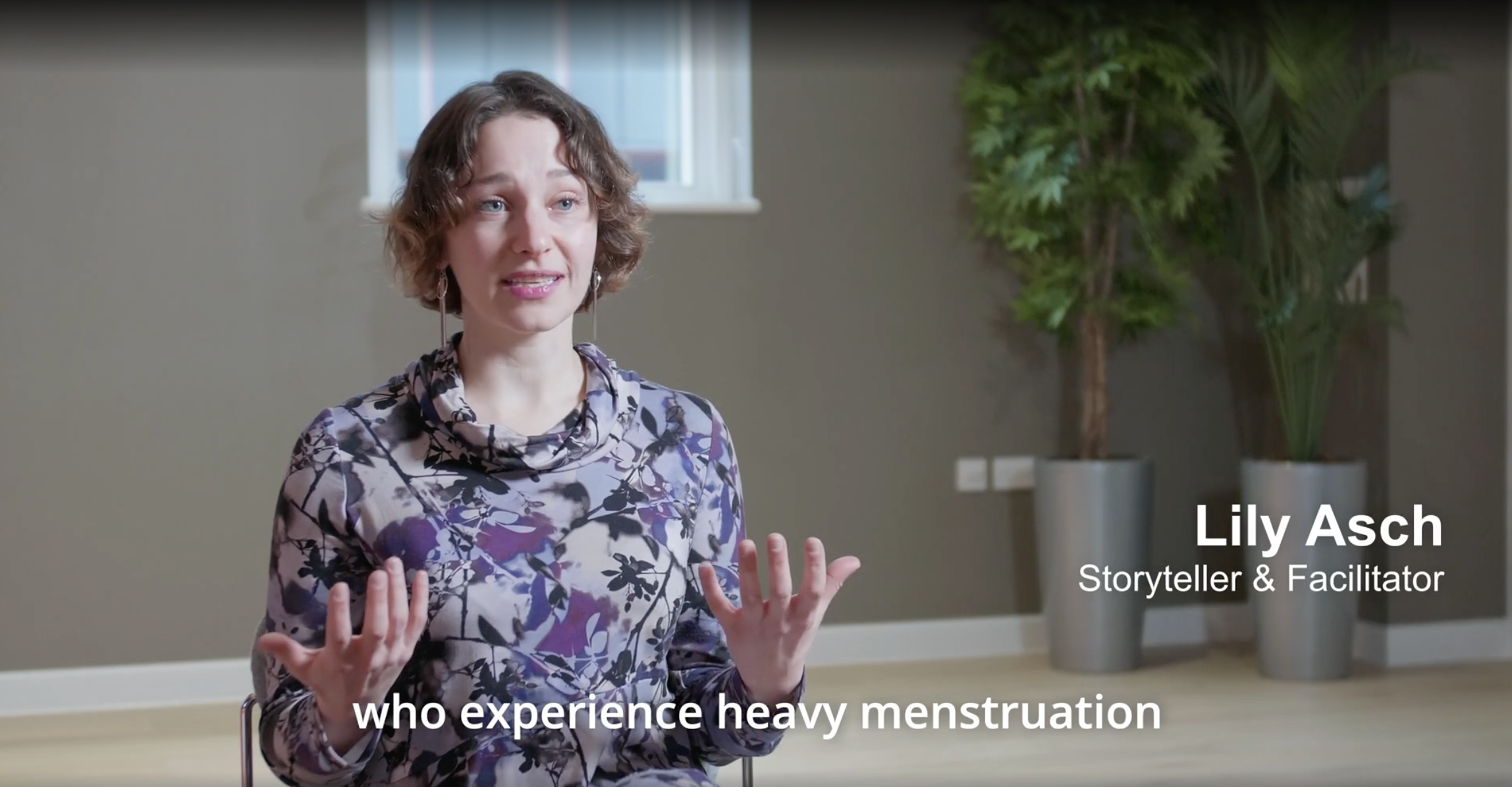 Lily Asch, a storyteller and facilitator, speaking during an interview or presentation indoors, with a beige wall, window, and large potted plants in the background, wearing a patterned top.