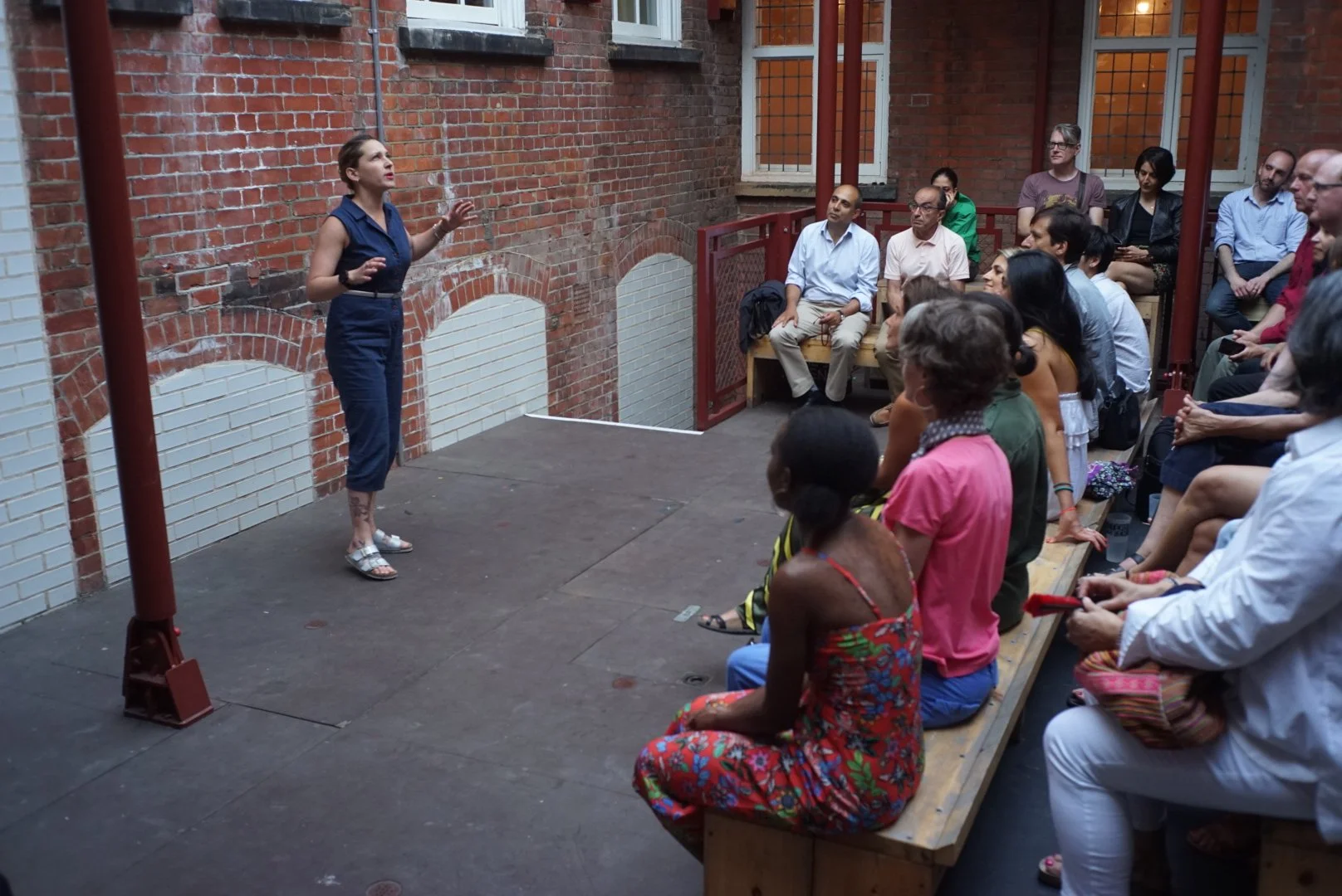 A woman is giving a talk to an audience sitting on benches and standing in a small outdoor patio with brick walls.