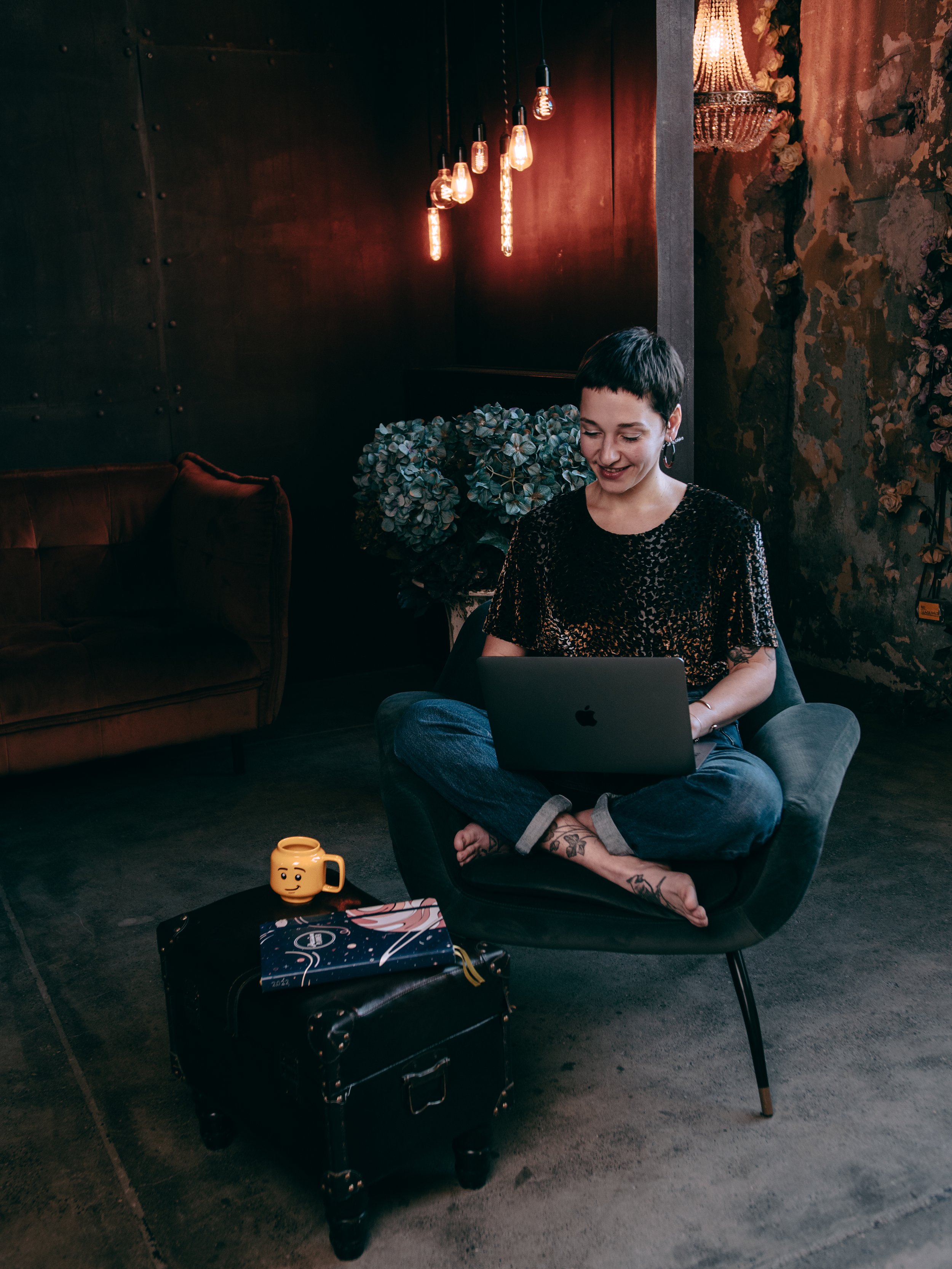 A woman with short dark hair and tattoos on her legs sitting cross-legged in a lounge chair, working on a silver laptop in a cozy, dimly-lit room with vintage everyone lighting and floral decor.