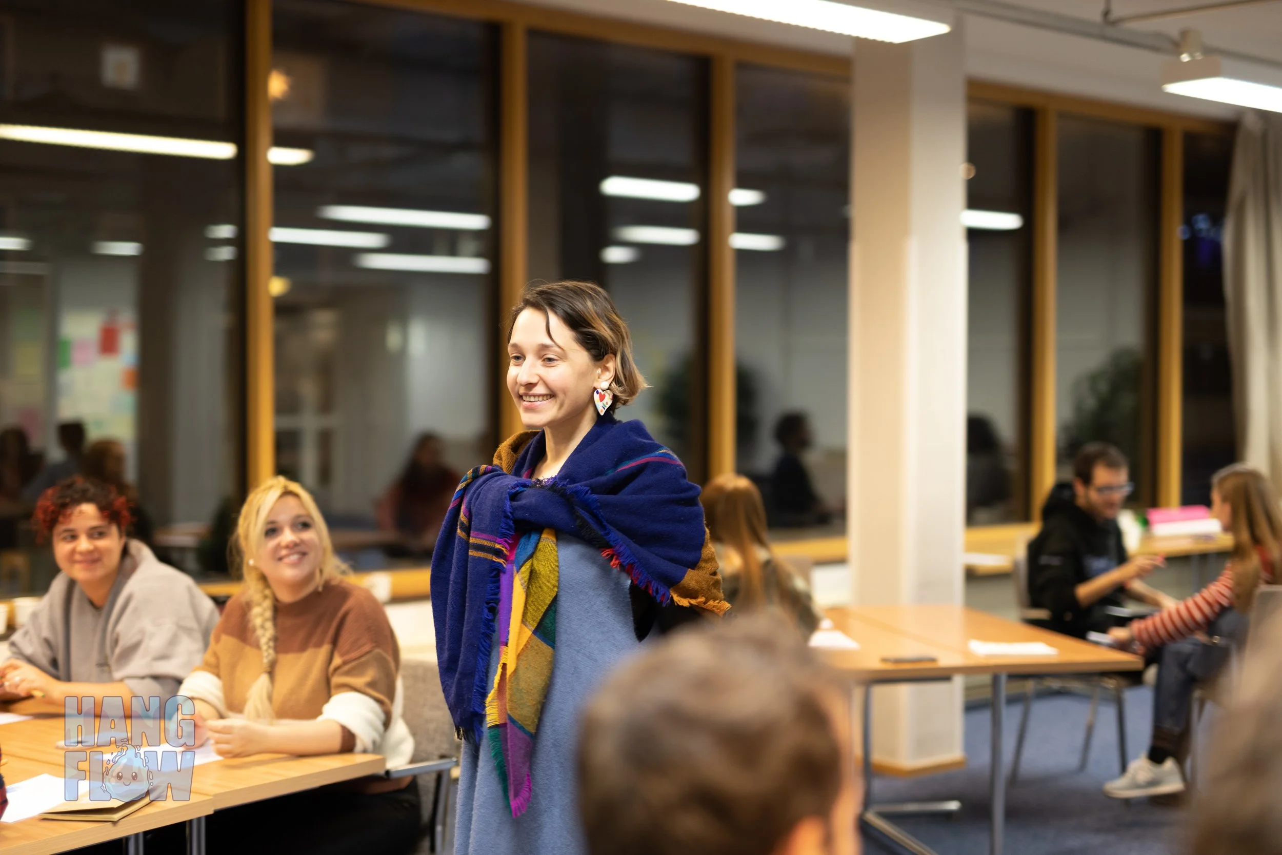 A group of diverse young adults in a classroom or study space, with one woman standing in the center smiling, wearing a colorful scarf and earrings, while others are seated around tables, engaging in conversation and studying.