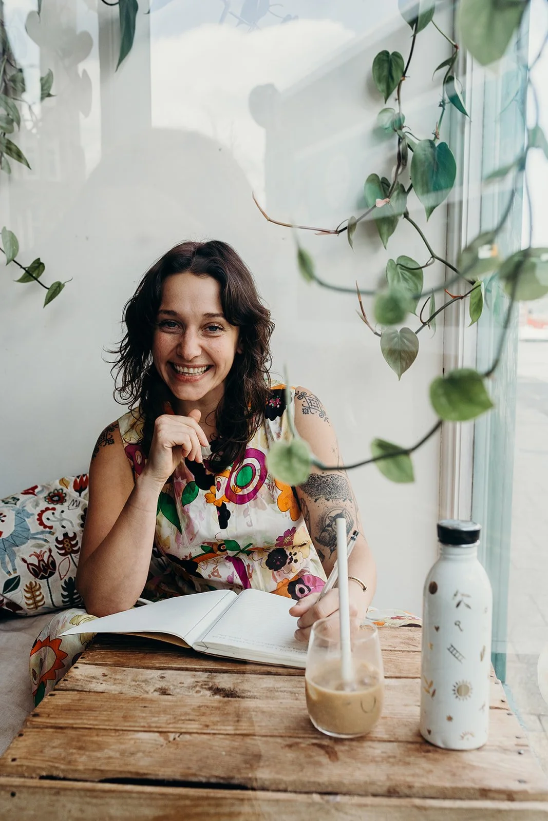 A woman with curly dark hair smiling and sitting at a wooden table in a bright cafe. She has tattoos on her arms and is wearing a colorful floral dress. On the table, there is an open notebook, a glass of iced coffee with a straw, and a decorated white water bottle. Green vines hang around a window behind her.