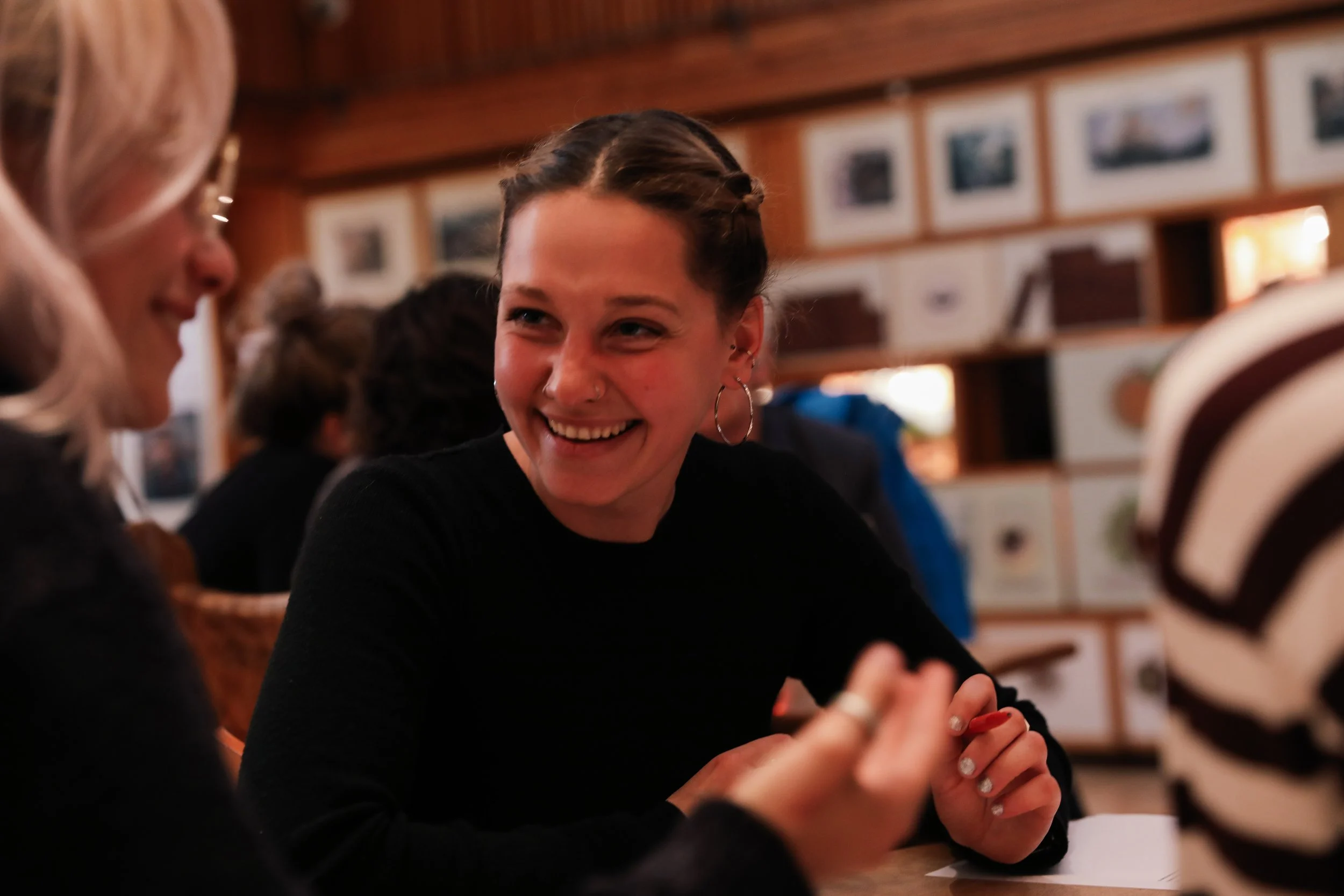A young woman with dark hair in braids, hoop earrings, smiling and talking to another woman with blonde hair in a setting with wooden walls and framed pictures.