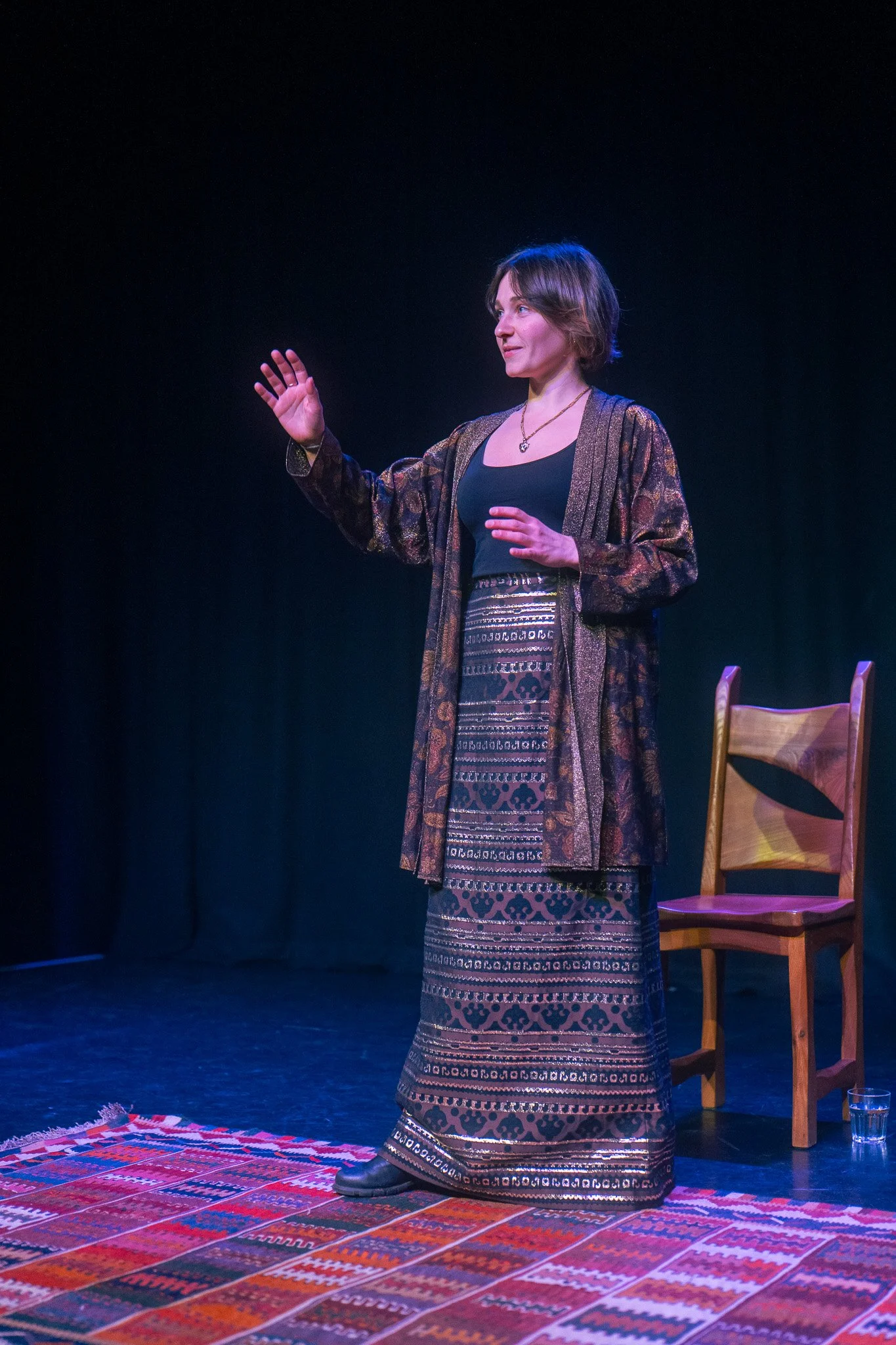 A woman performing on stage with a black background, standing on a colorful patterned rug, wearing a long skirt, black top, and a patterned coat, with a wooden chair and a glass of water behind her.