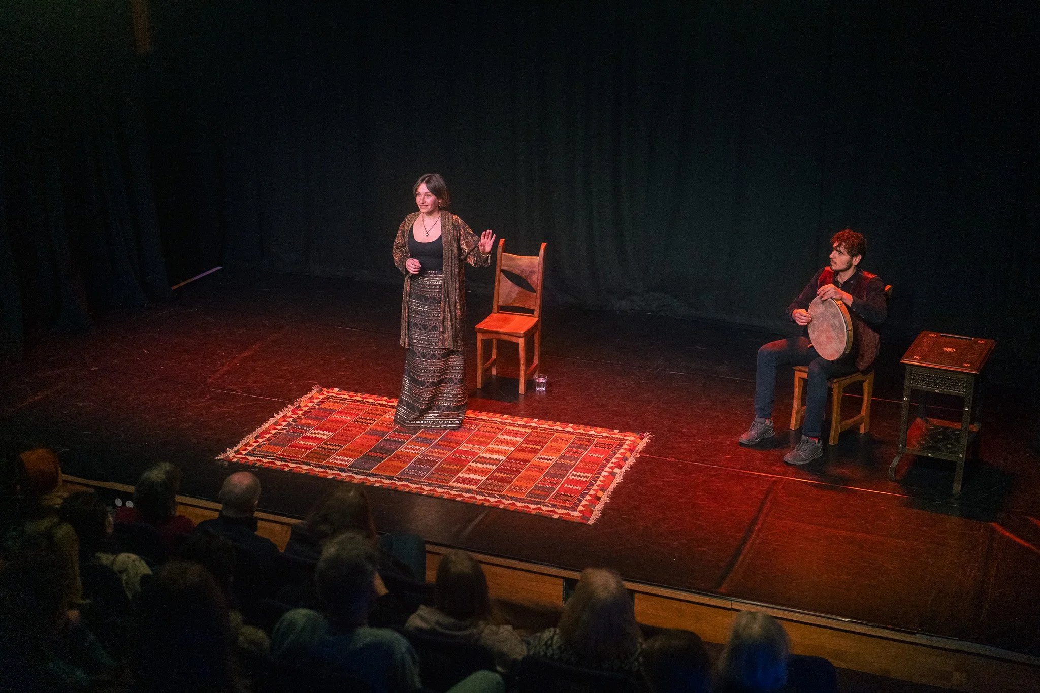 Lily Asch stands on stage speaking to an audience, with a man seated behind her playing a drum. The stage has a colorful rug, two chairs, and a small table. The background is black, and the audience is visible in the foreground.