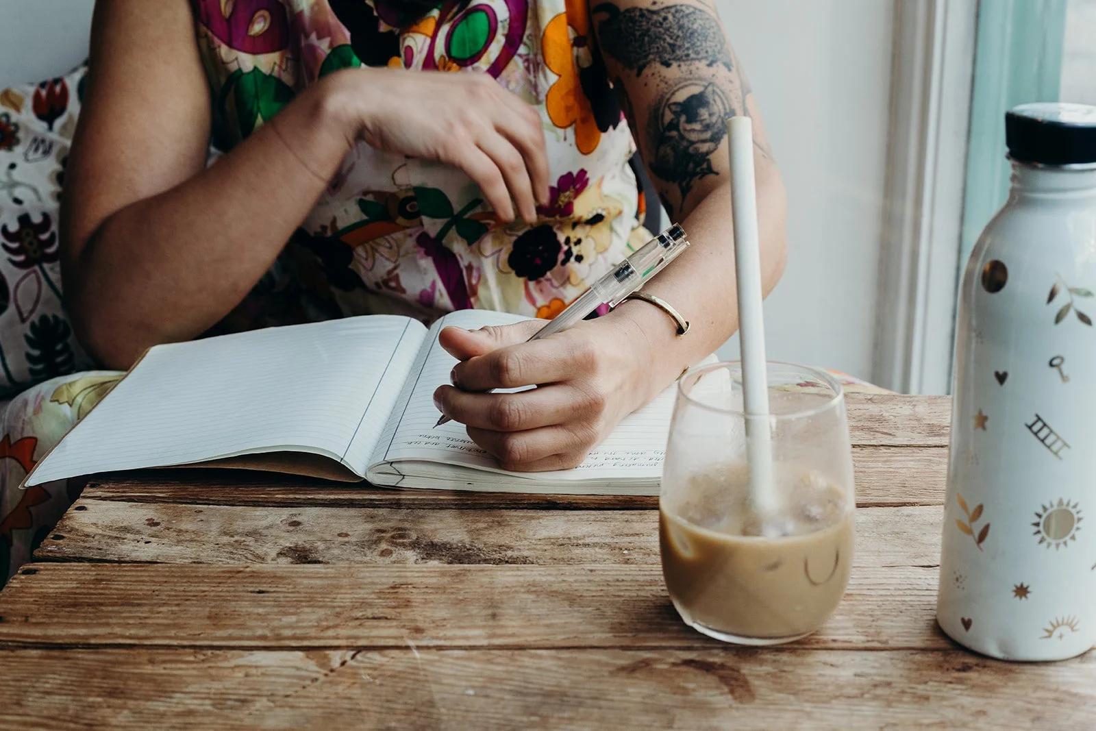 A person with tattoos on their arms is sitting at a rustic wooden table, writing in a notebook with a pen. There is a glass with iced coffee and a white bottle decorated with celestial patterns on the table, near a window with a curtain.