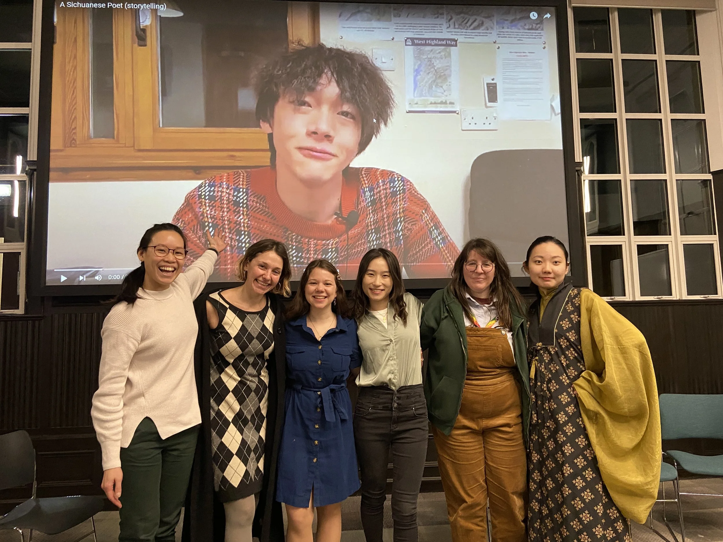 Group of six women standing in front of a large screen displaying a young man with curly hair. The women are smiling and posing together.